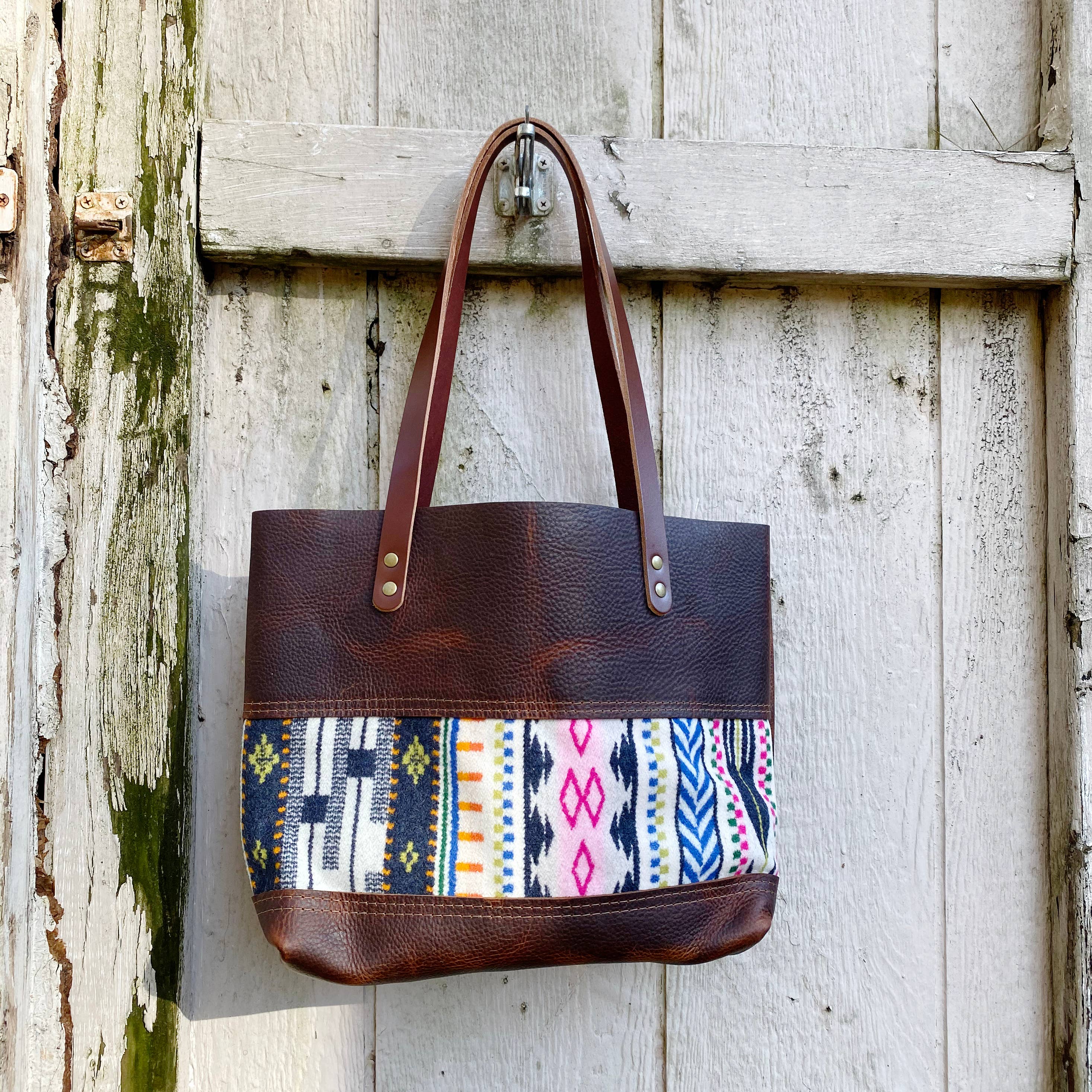 The Hawks & Doves Wool Tote Short is a brown full grain leather tote with two handles and a vibrant, patterned fabric panel in pink, blue, yellow, and white inspired by Pendleton wool designs.