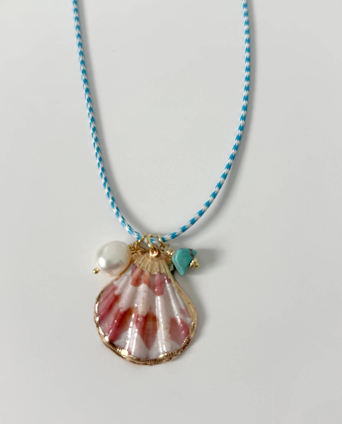 The Islas Necklace by Tramps+Thieves showcases a blue-and-white braided cord, a striking pink-and-white seashell pendant, a turquoise bead, and a white pearl charm—perfect for ocean-inspired style.