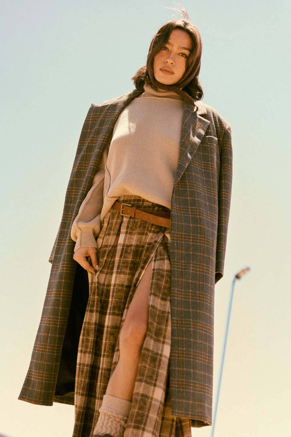 A woman wears the Promesa USA Plaid Felt Double-Breasted Calf-Length Overcoat, along with a beige turtleneck, high-slit plaid skirt, and socks, posing confidently outdoors against a clear sky.