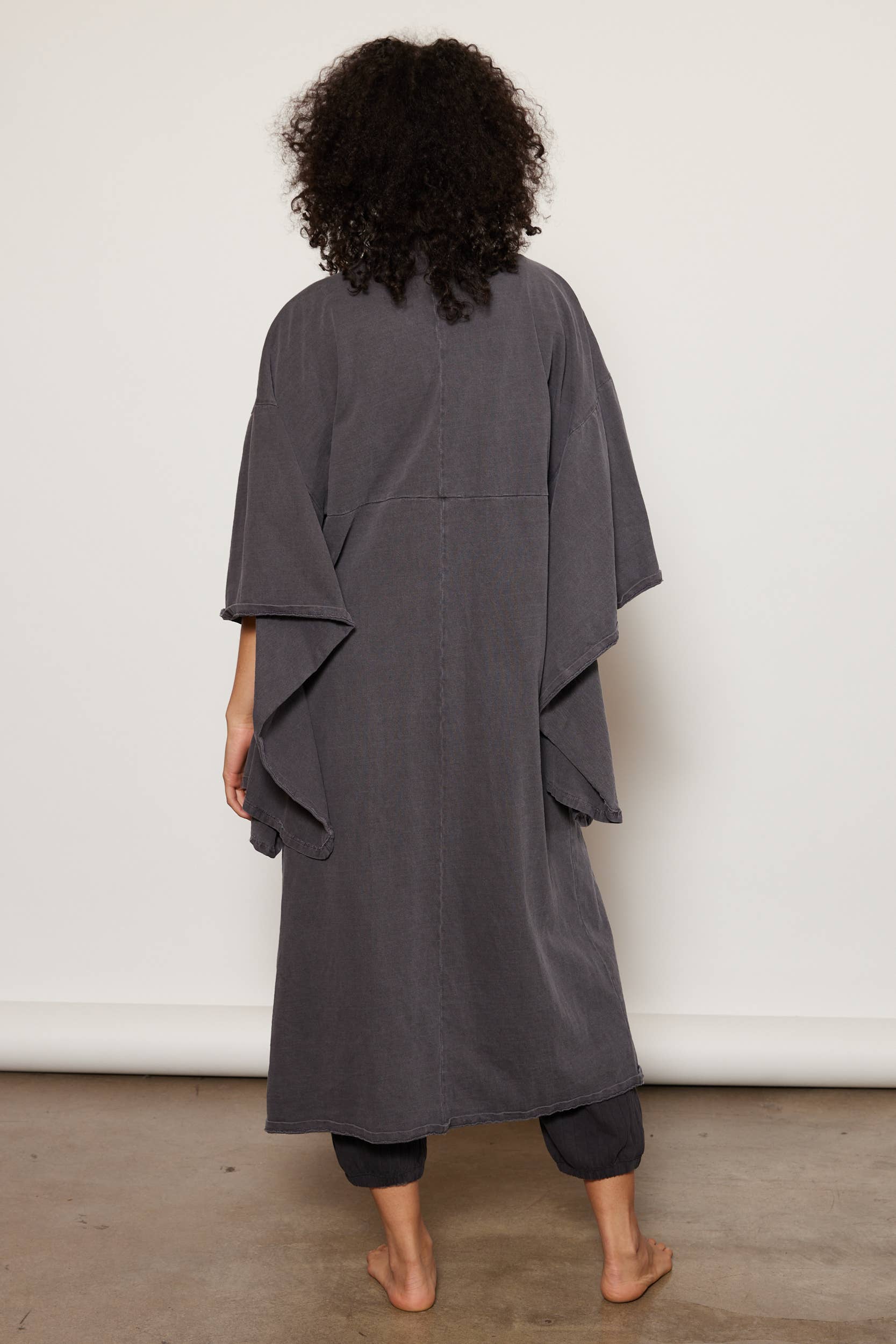 A person with curly hair stands barefoot, facing away, wearing People Of Leisures loose, dark gray organic cotton Dawn Robe over matching pants. The setting features a light wall and concrete floor.
