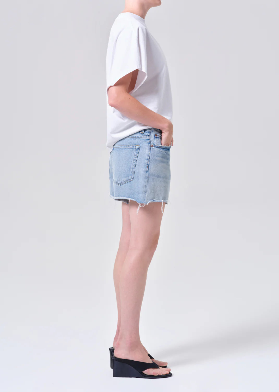 Model wearing NYE SHORT in post, light blue denim with raw hem, paired with white tee and black wedge sandals