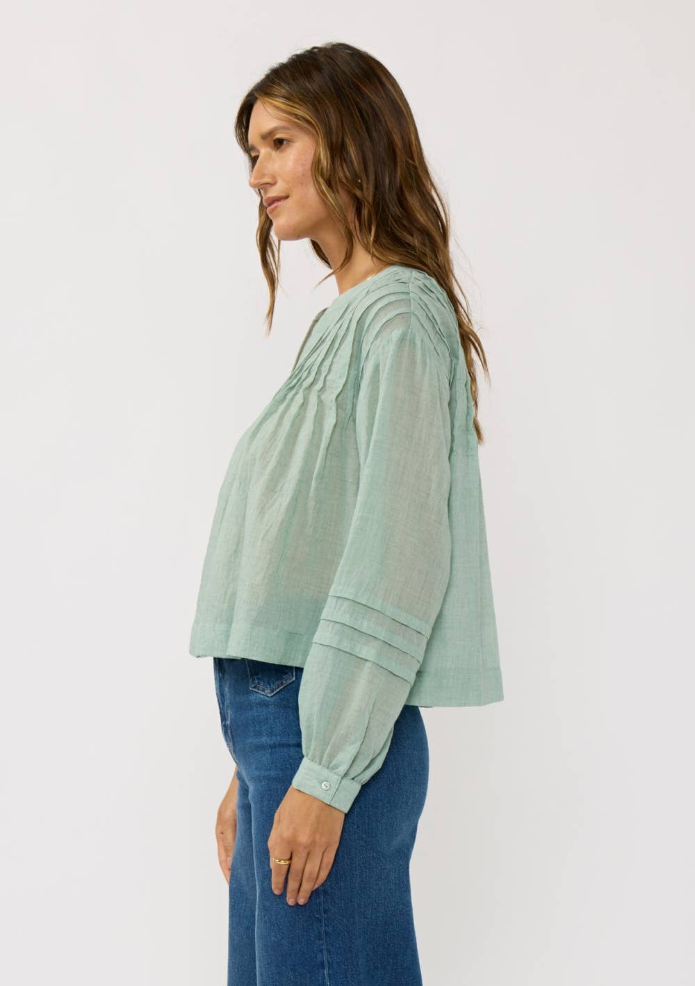 Woman wearing a light green Pintuck Pleated Button-Down Blouse with long sleeves paired with blue jeans standing sideways