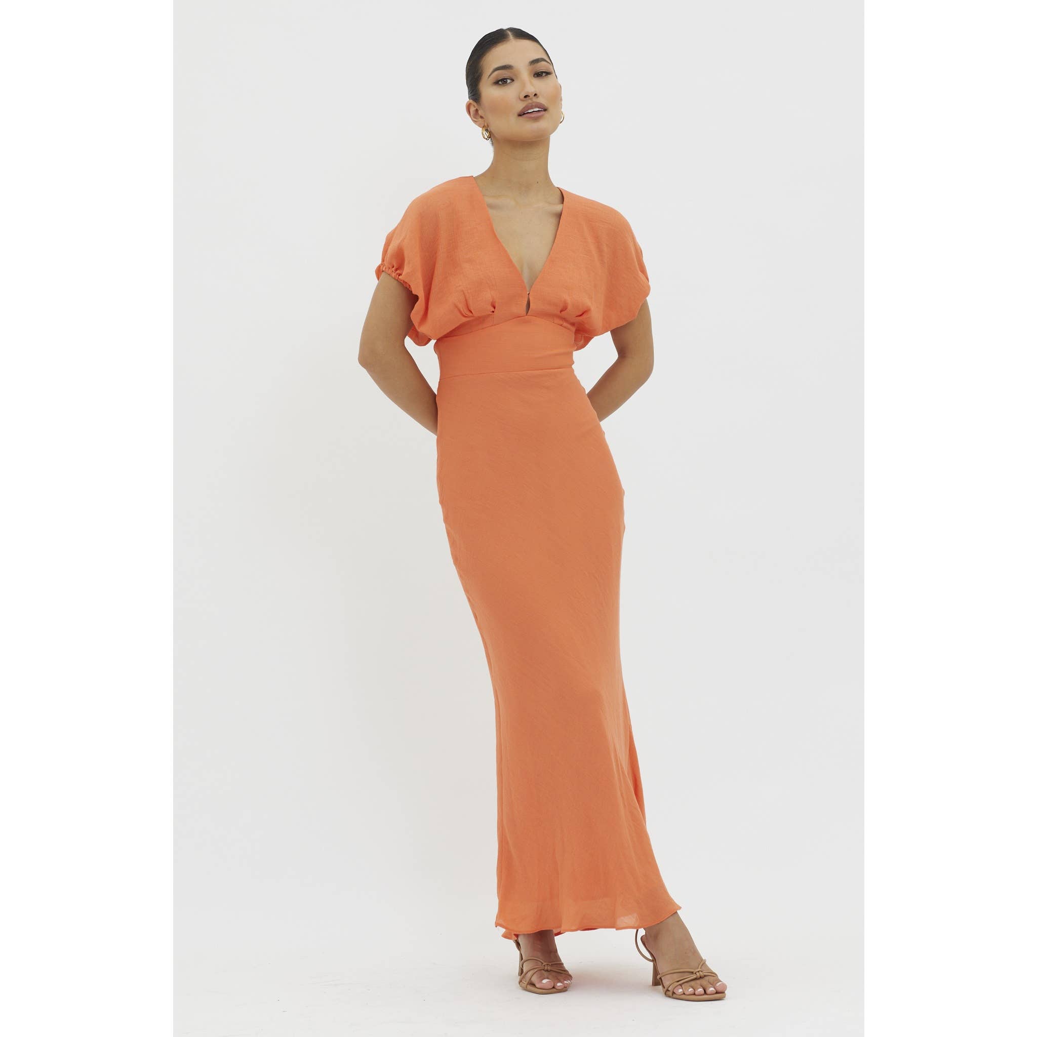 A woman poses against a white background wearing the One and Only Collective Tangerine V-neck Maxi Dress, featuring short sleeves, an elastic waistband, floor-length orange fabric, and strappy heels. Her hair is pulled back.