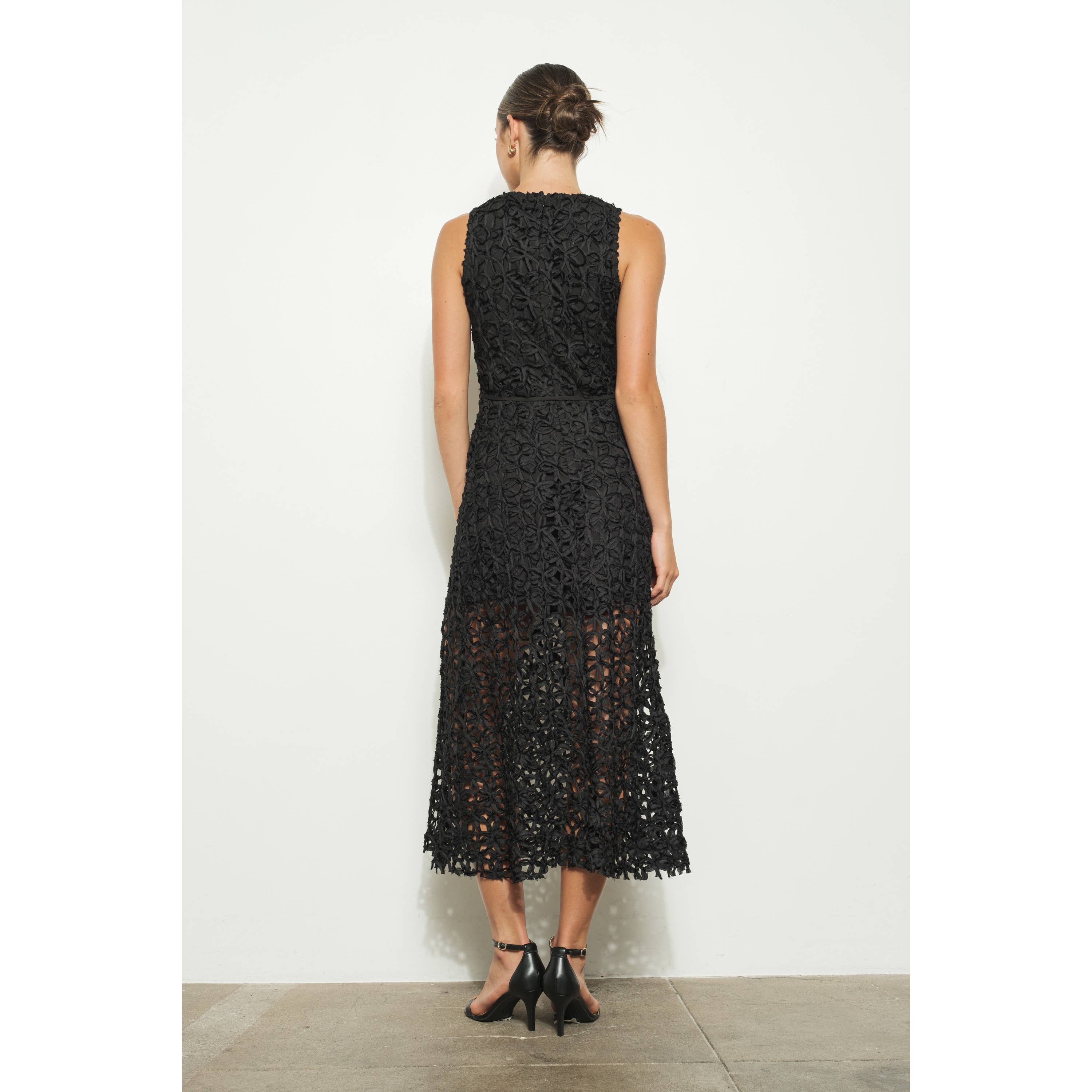 A woman in a neat bun poses with her back to the camera against a plain white wall, wearing the INA Sleeveless Crochet Lace Overlay Midi Dress with a sheer midi skirt and black high-heeled sandals.