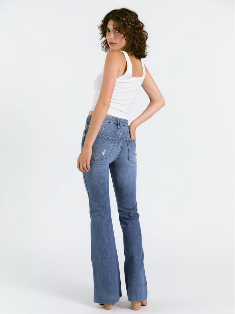 A woman with curly hair models Porter Blue Apparel’s WANDERER 5 POCKET FLARE IN MABEL WASH, featuring a high-rise waistband, paired with a white tank top as she stands and looks over her shoulder against a white background.