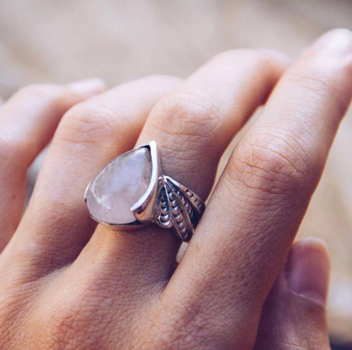 A close-up of a hand wearing the bahgsu jewels Winged Ring, featuring a large, teardrop moonstone set in an ornate silver band. The softly blurred background highlights its shimmering gemstone and intricate craftsmanship.