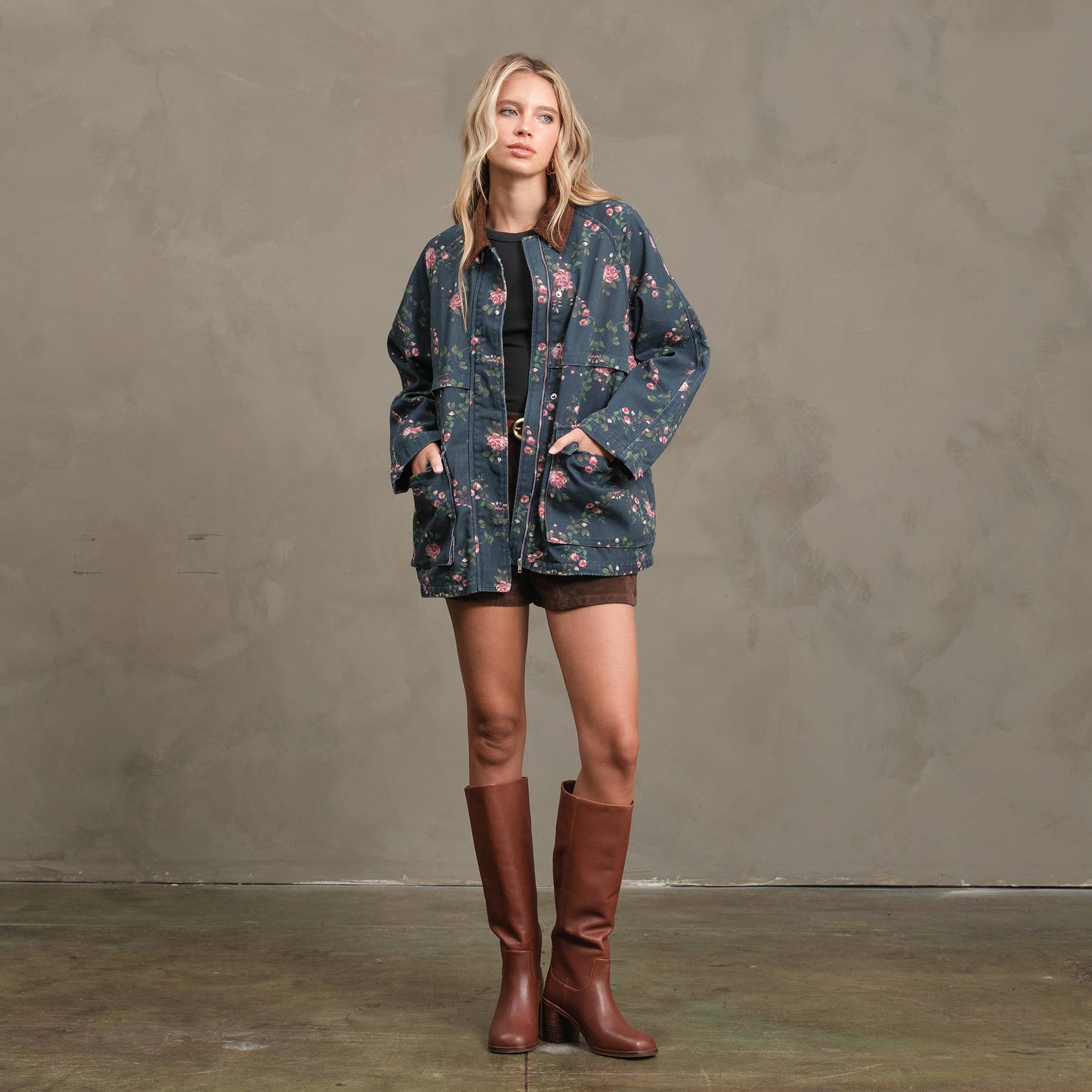 A woman stands against a textured wall wearing the RAISONNEL Floral Printed Utility Jacket (RJ1073), black shorts, and knee-high brown boots, hands in her pockets and a relaxed expression on her face.