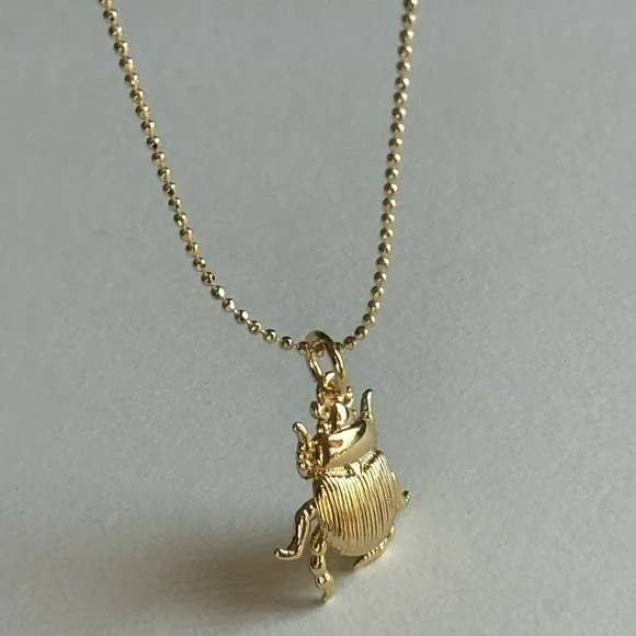 The Tramps+Thieves Scarab Necklace features a small gold-filled beetle pendant on a delicate beaded chain, resting elegantly on a light, neutral surface.