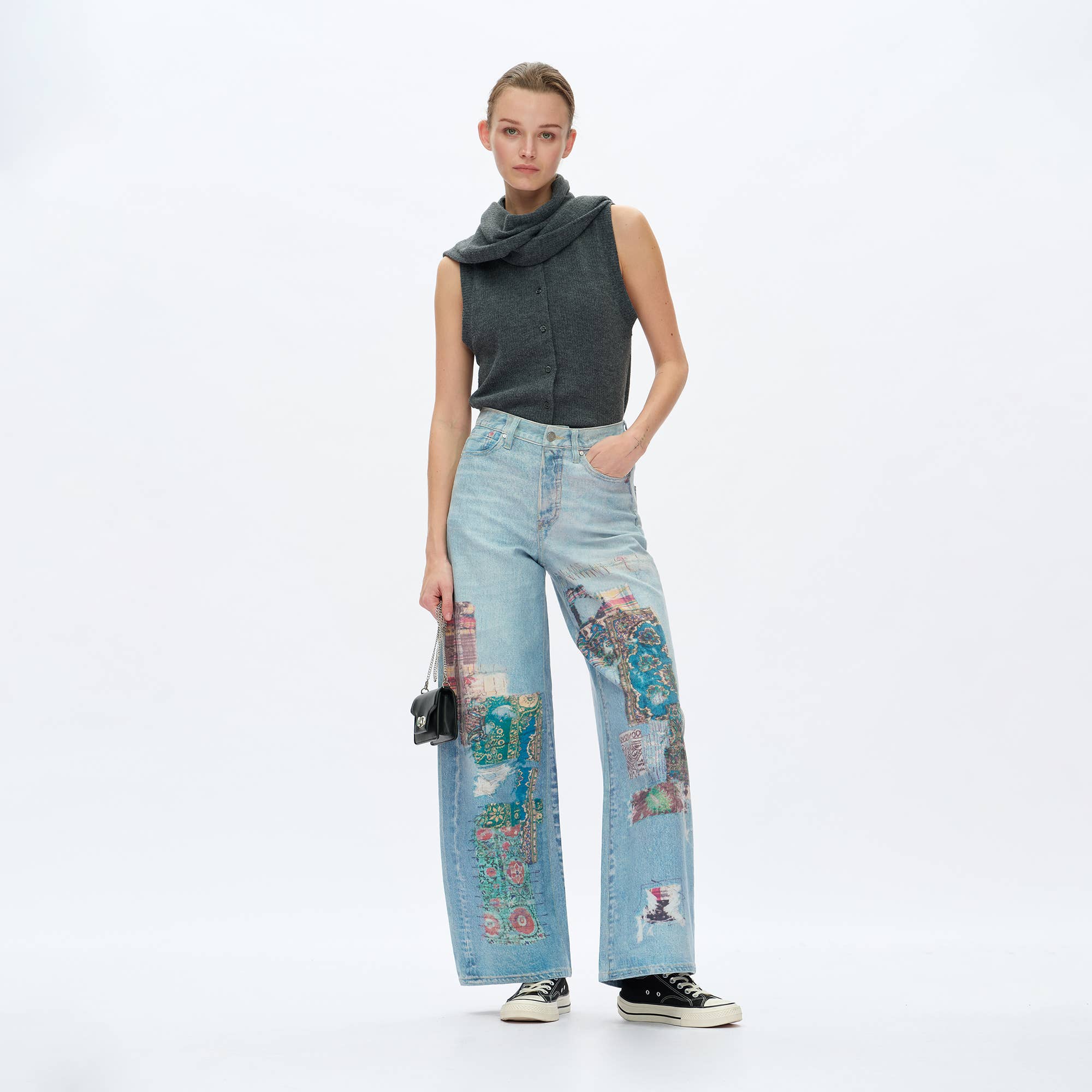 A woman poses against a white backdrop wearing BAYEAS Light Blue Patch Pattern High-Rise Barrel Jeans, a sleeveless gray cowl-neck top, black Converse sneakers, and holding a small black handbag.