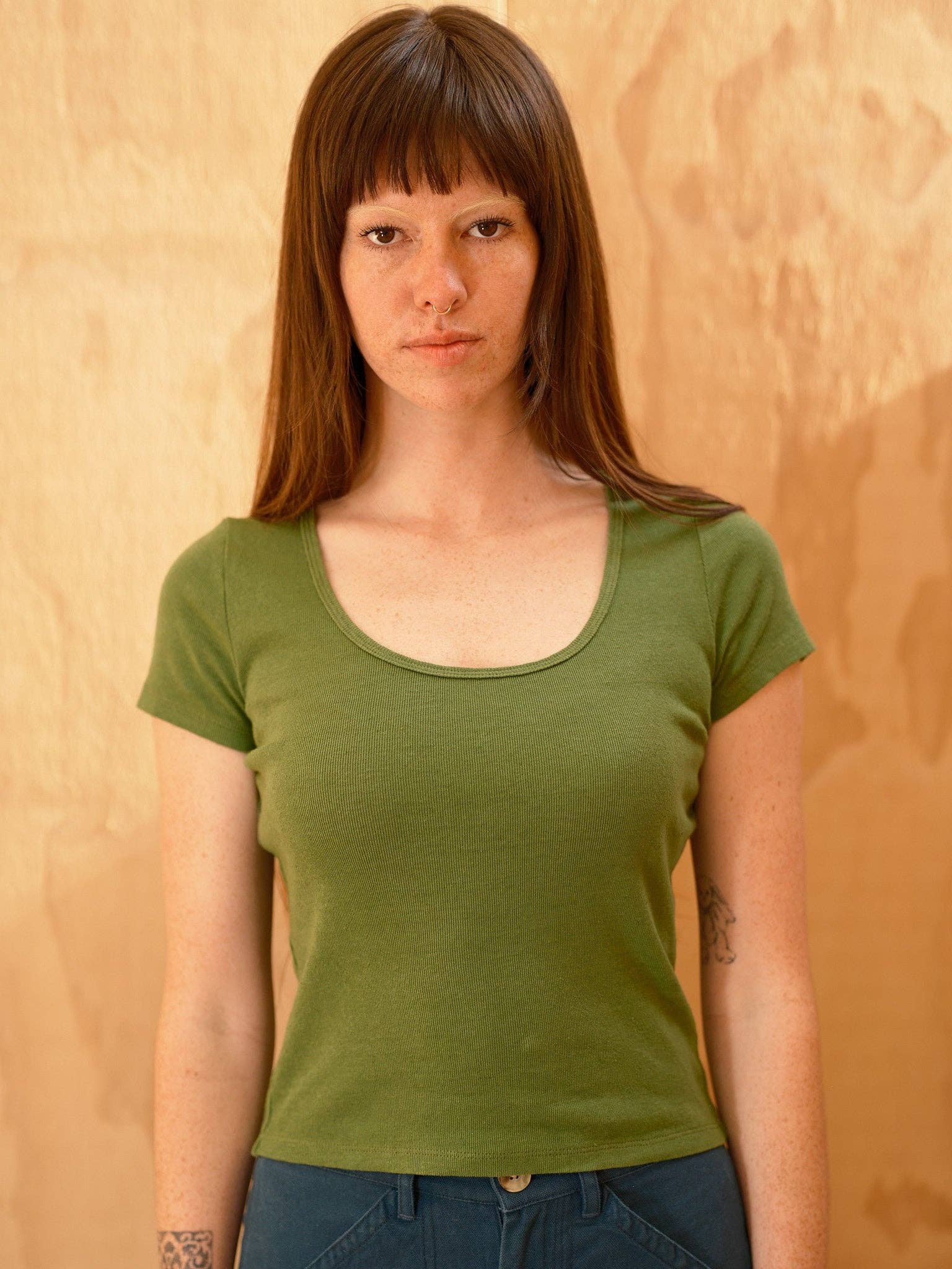 A person with long brown hair and bangs wears the Mollusk Scoop Ribsy Tee in green and blue pants, standing against a warm beige textured wall and looking directly at the camera with a neutral expression.
