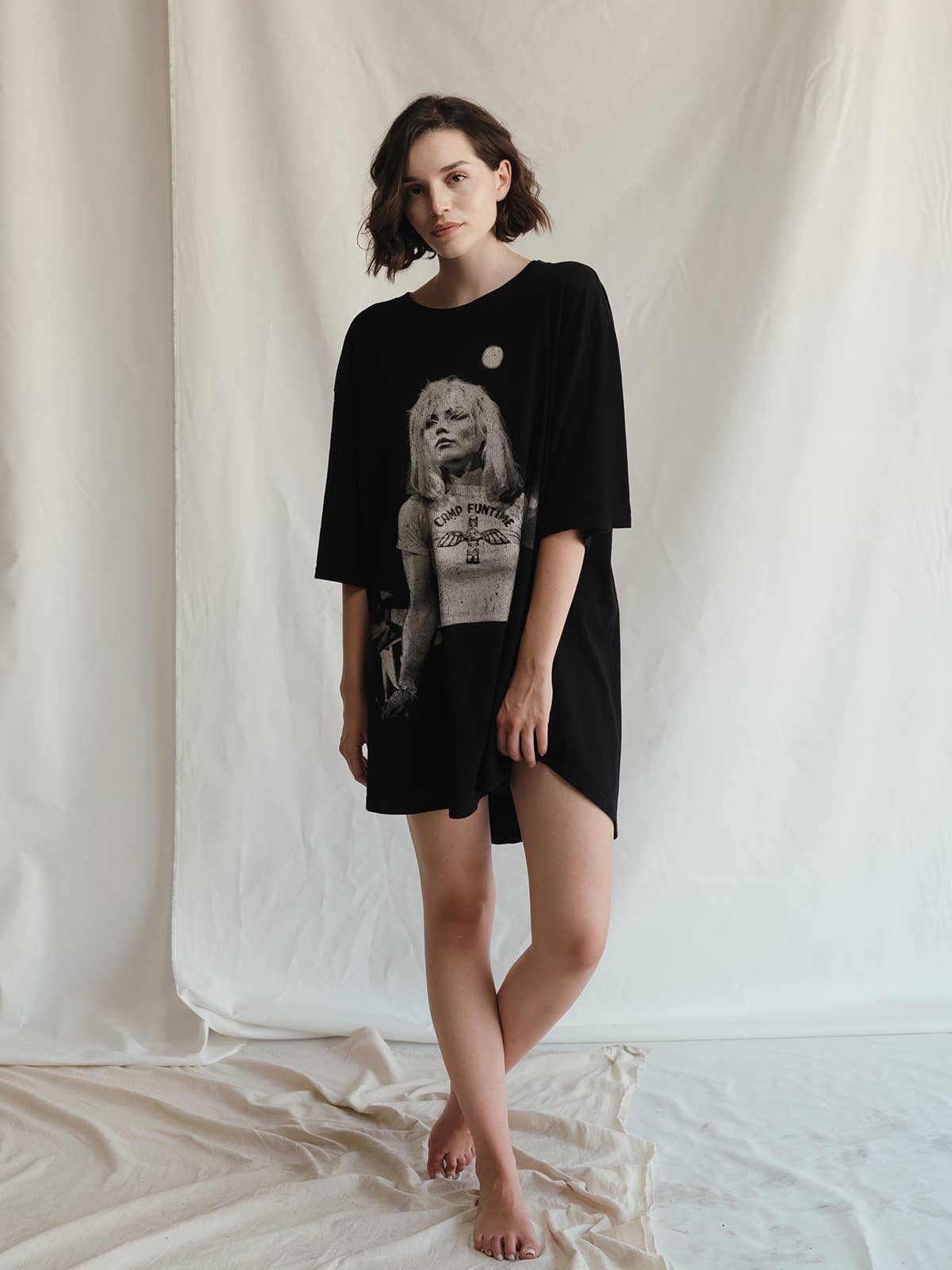 A young woman with short brown hair stands barefoot on a white draped floor, wearing the People Of Leisure Blondie Camp Oversized Tee—an ethically made black shirt with a graphic print of a blonde woman—set against a plain, light fabric backdrop.
