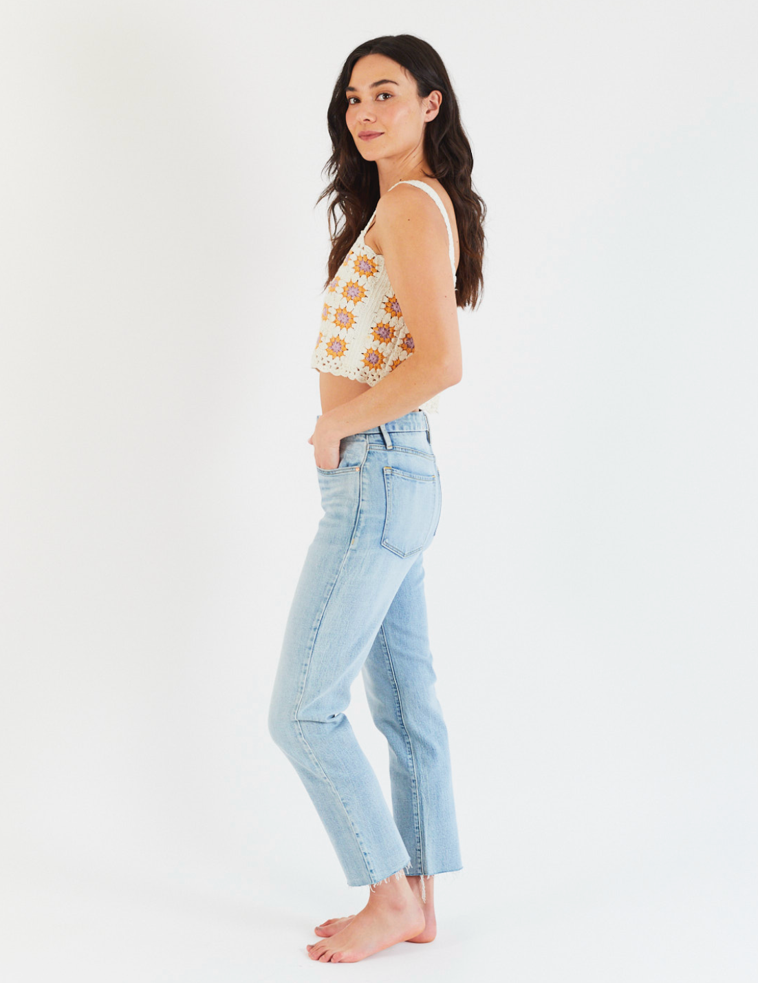 A woman stands barefoot in Porter Blue Apparels REBEL STRAIGHT IN EDEN WASH jeans and a sleeveless floral crop top, facing sideways with her head turned toward the camera, set against a plain white background.