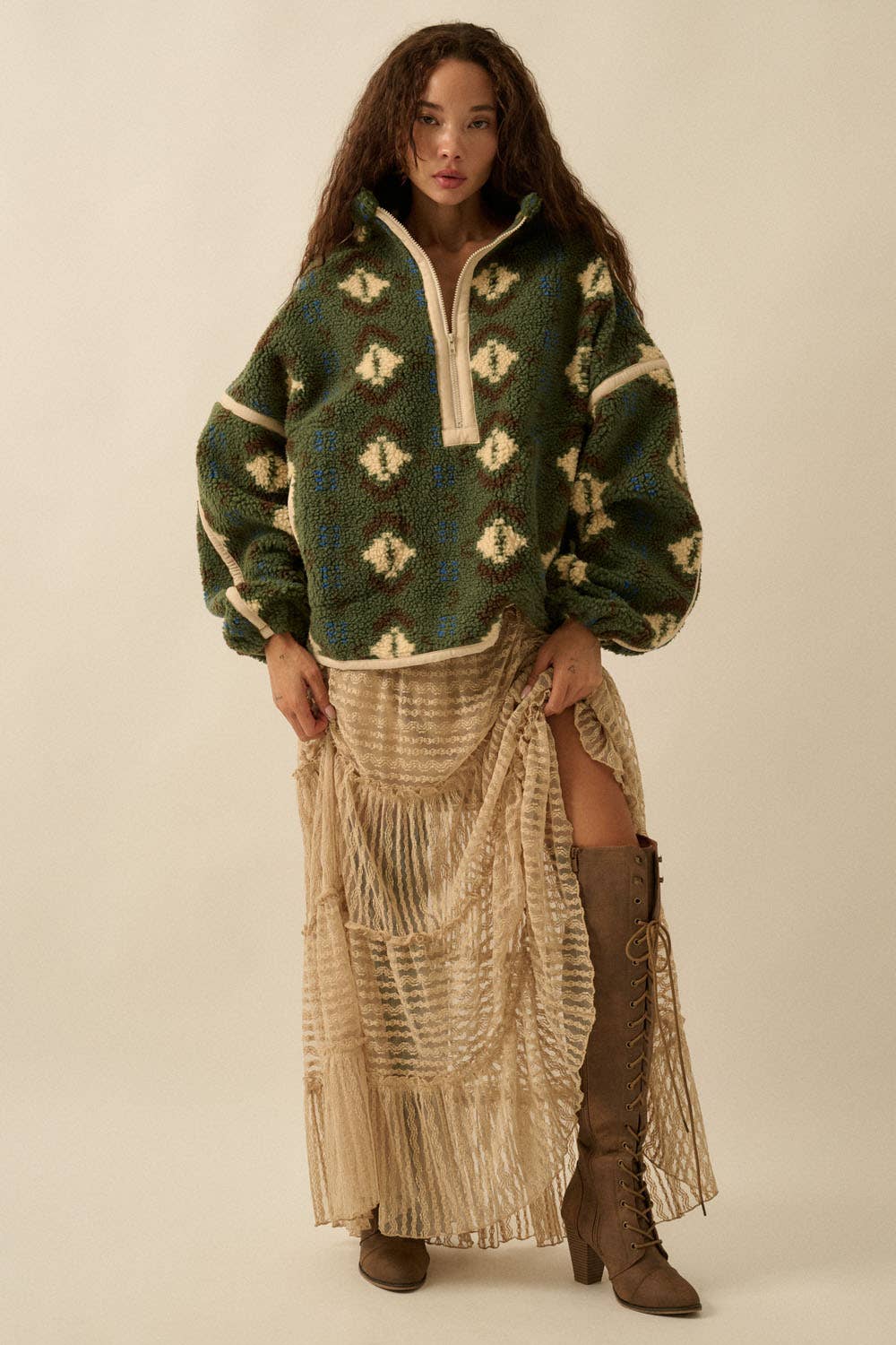 A woman with long curly hair wears the Promesa USA Geo-Print Sherpa Fleece Half-Zip Pullover Jacket, a beige fringed skirt with a slit, and knee-high brown lace-up boots, standing against a neutral background.
