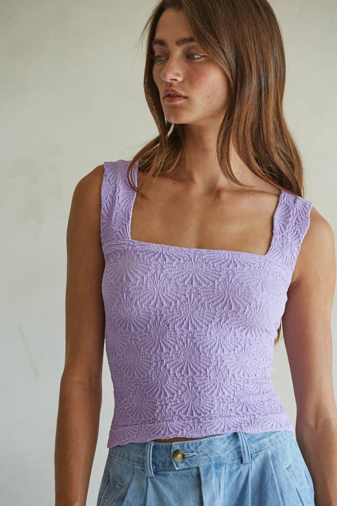 A woman with long brown hair wears the By Together Knit Modal Nylon Spandex Seamless Jacquard Tank in lilac, featuring a square neckline, paired with high-waisted light blue pants. She looks to the side against a neutral background.