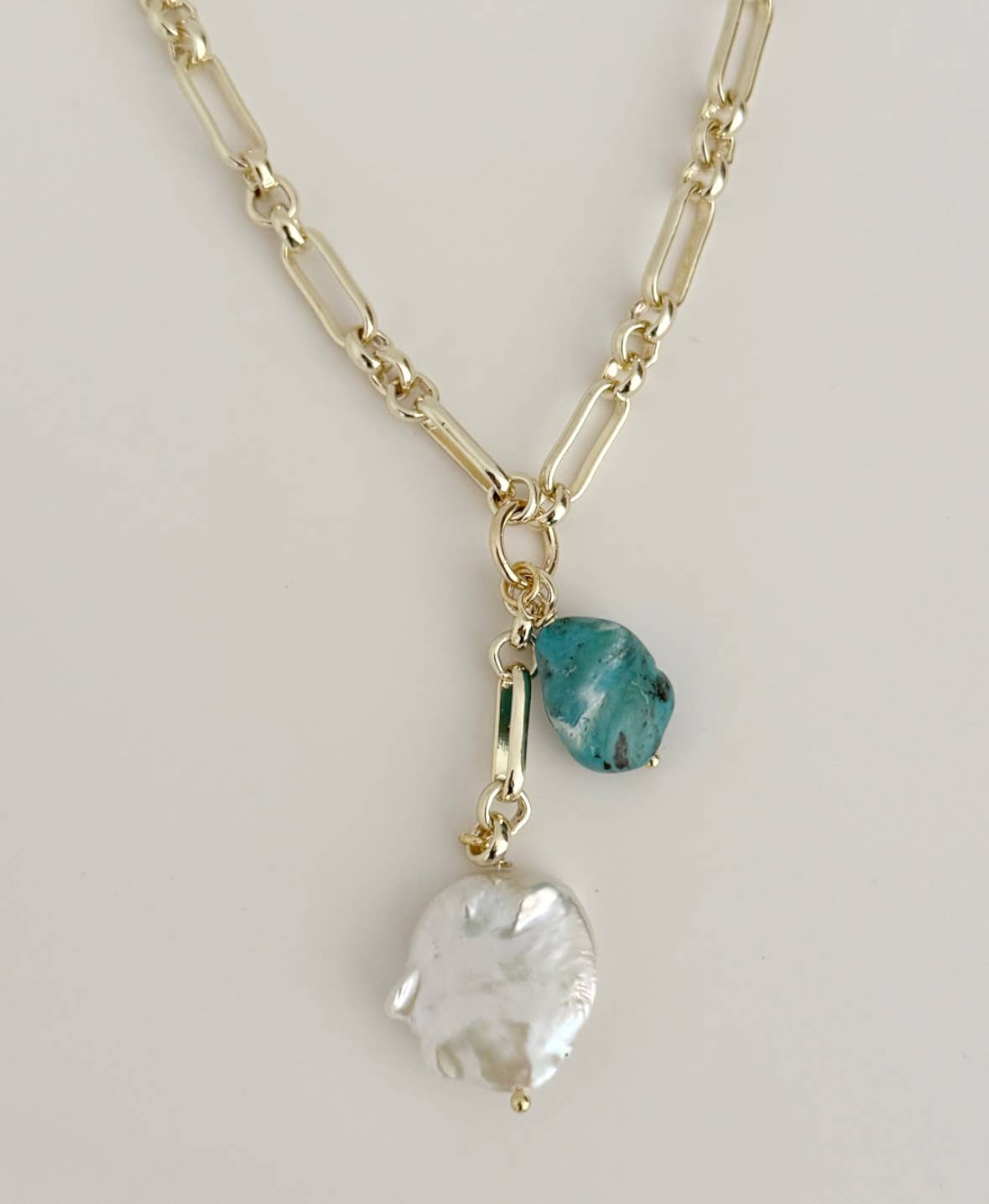 The Azure Tide Necklace by Tramps+Thieves features an 18K gold-filled chain with three pendants: a baroque pearl, rough blue stone charm, and a small rectangular green gem, all elegantly suspended together.