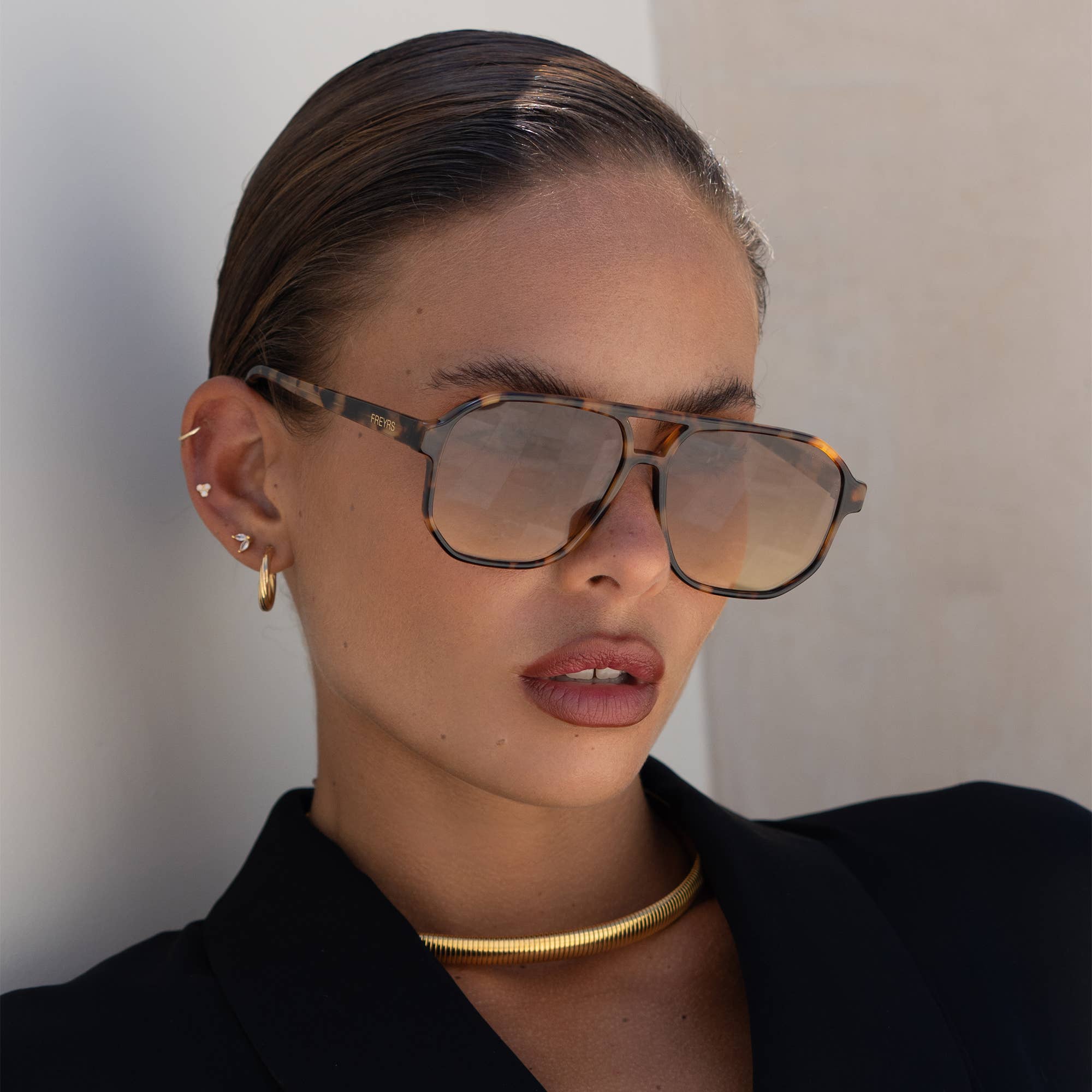 A woman with slicked-back hair wears FREYRS Eyewears Billie (NEW) unisex aviator sunglasses with premium nylon lenses, gold jewelry, and a black blazer, posing against a light background.