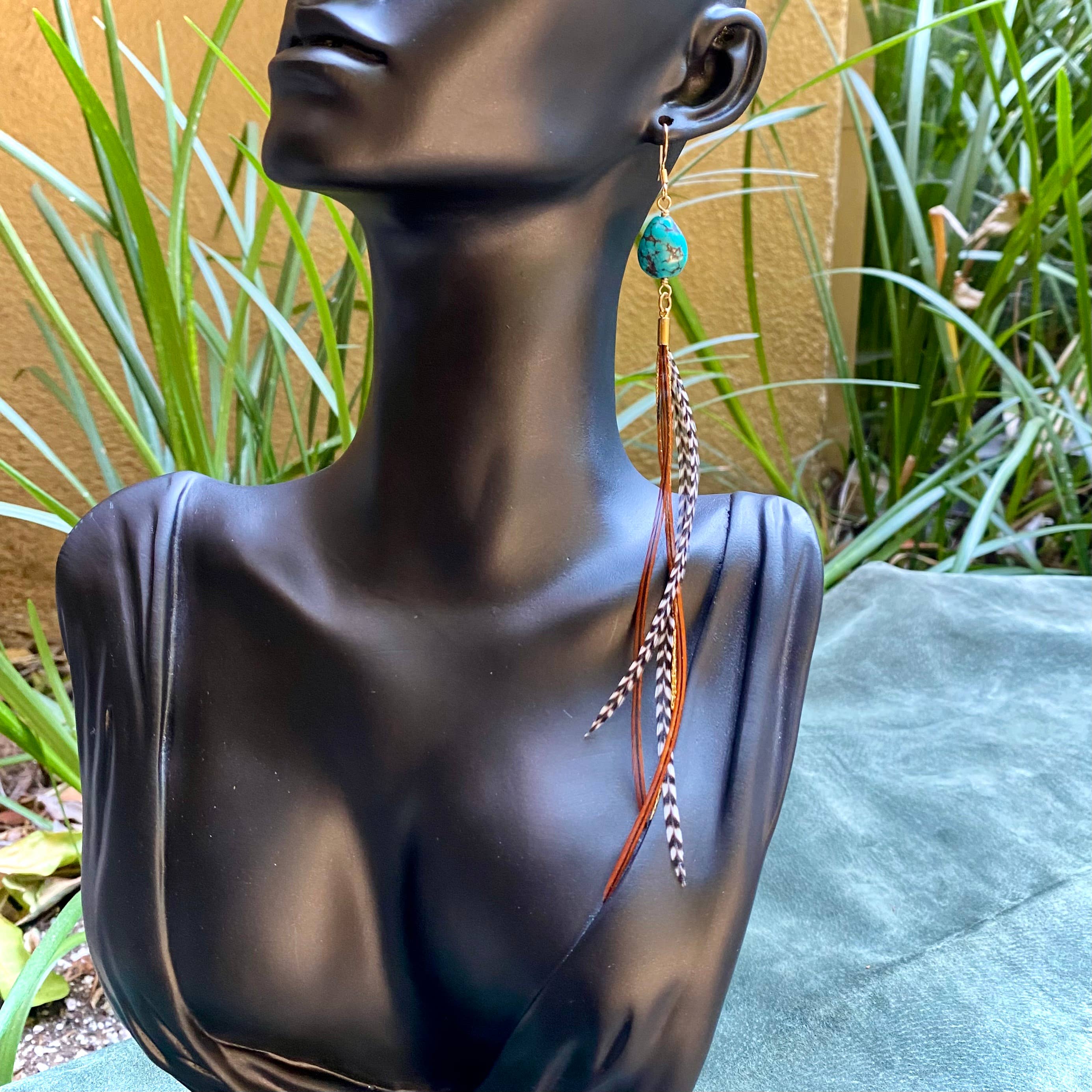 A black bust displays ASTALI’s Turquoise & Mini Feather Earrings – Grizzly & Fawn/Gold against a backdrop of green plants and a yellow wall. The earrings feature turquoise stone and mini brown and black-and-white striped feathers.