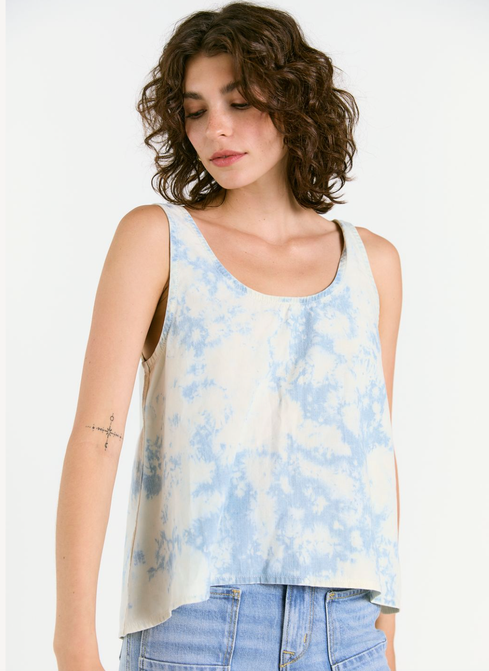 A person with curly brown hair wears the WABI SABI TANK IN SKY TIE DYE by Porter Blue Apparel, paired with light blue jeans. They have a small, minimalist compass tattoo on their upper left arm and look down with a neutral expression.