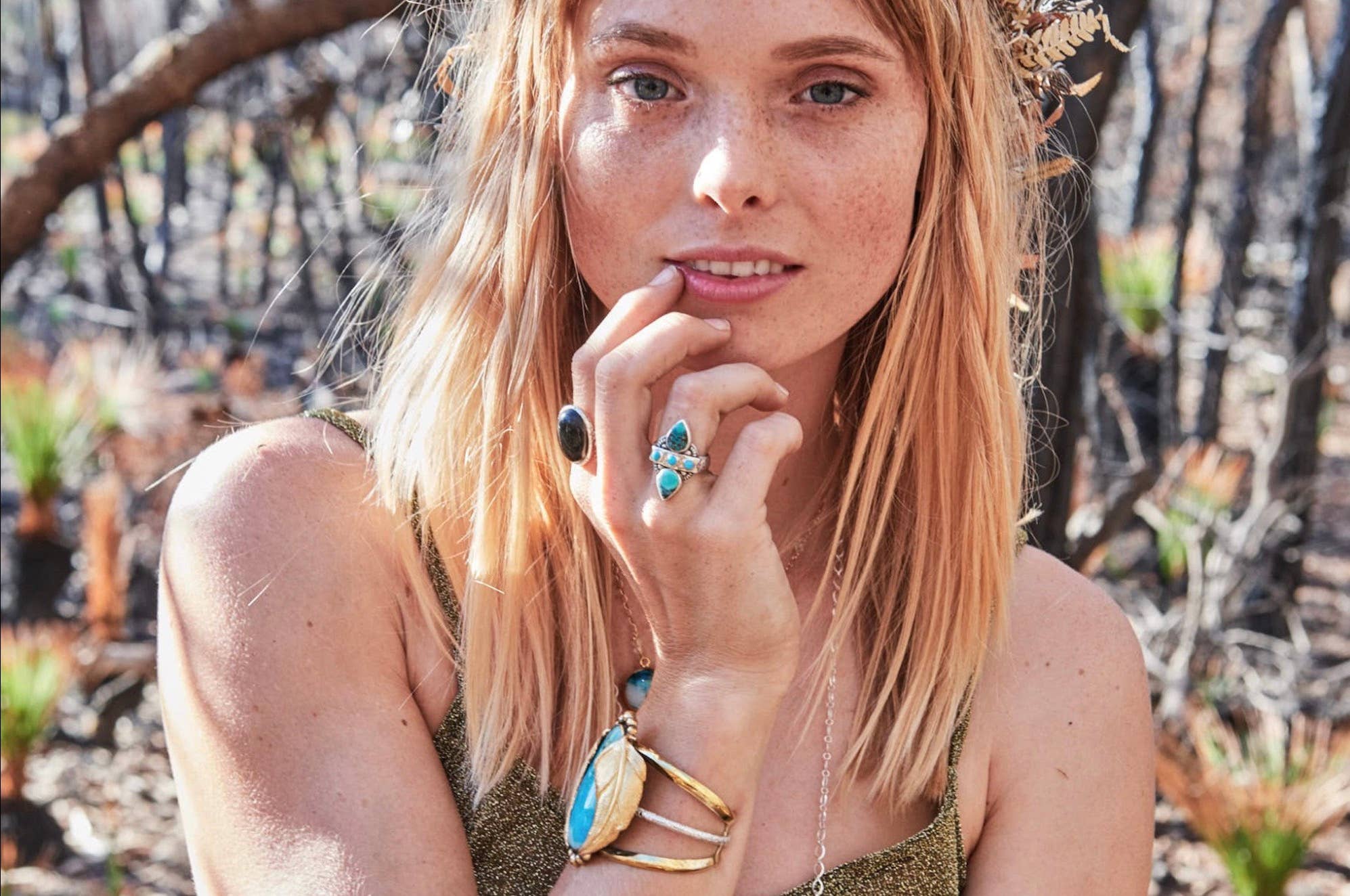 A light-skinned, blonde woman wears bahgsu jewels’ Truth Seeker Ring along with gold and turquoise jewelry. She stands in a sunlit wooded area, touching her chin and gazing at the camera.