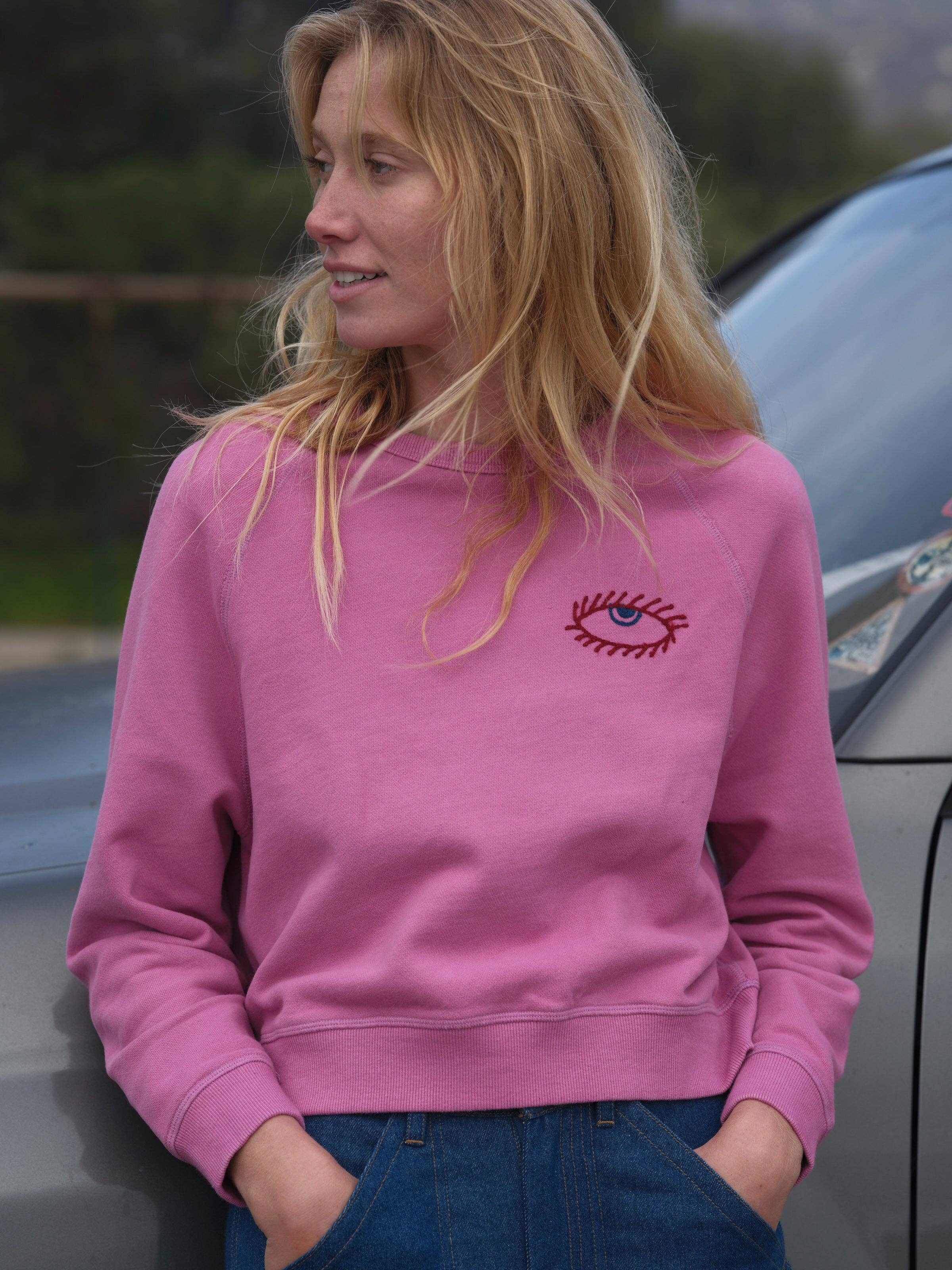 A woman with long blonde hair wears the Mollusk Visions Crew in pink, featuring an embroidered eye on the chest, paired with blue jeans. She stands outdoors beside a gray car, looking to her left and smiling gently.