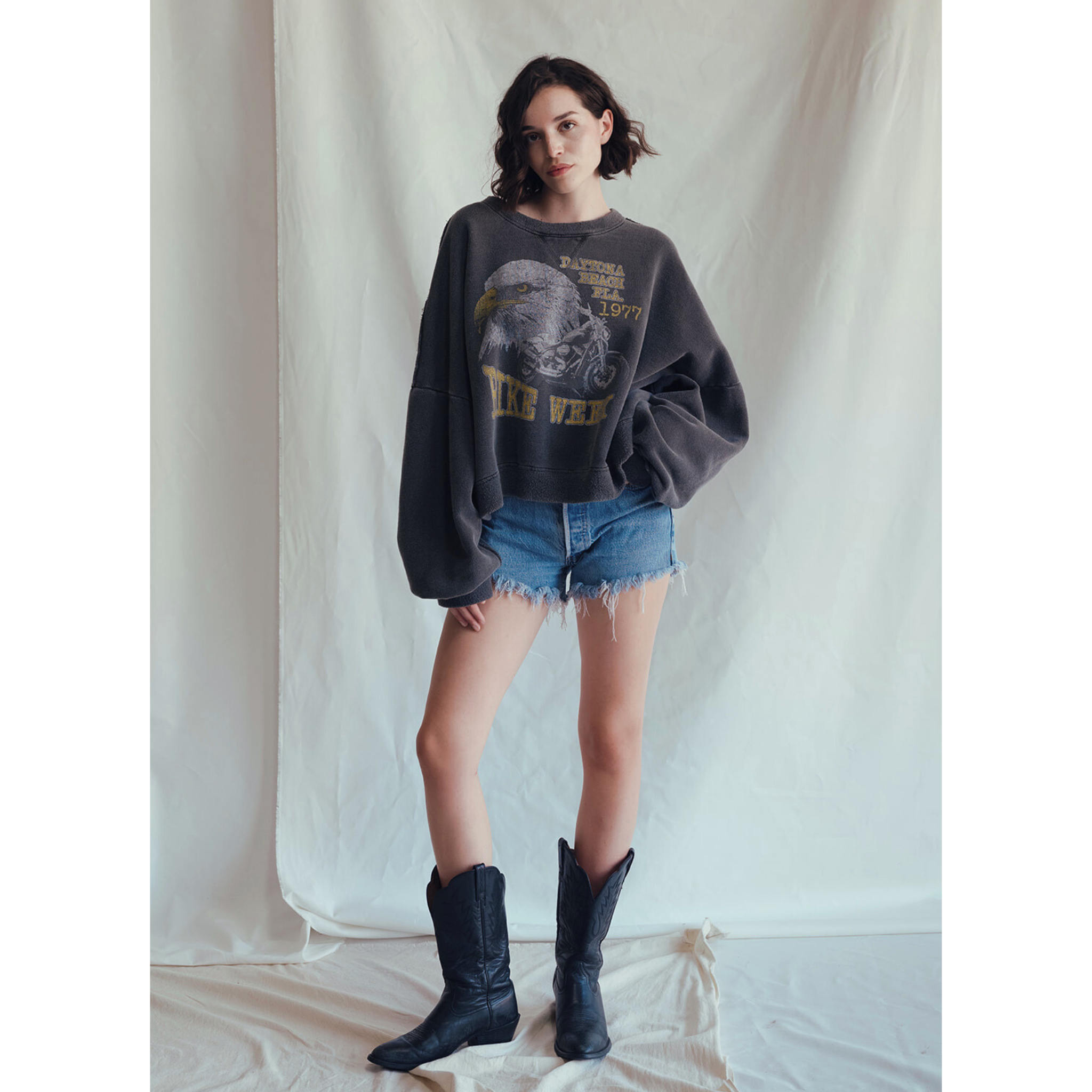 A woman with short, wavy dark hair stands in front of a light fabric backdrop, wearing the People Of Leisure Daytona Sweatshirt featuring a vintage eagle graphic, paired with denim shorts and black cowboy boots in a relaxed pose.