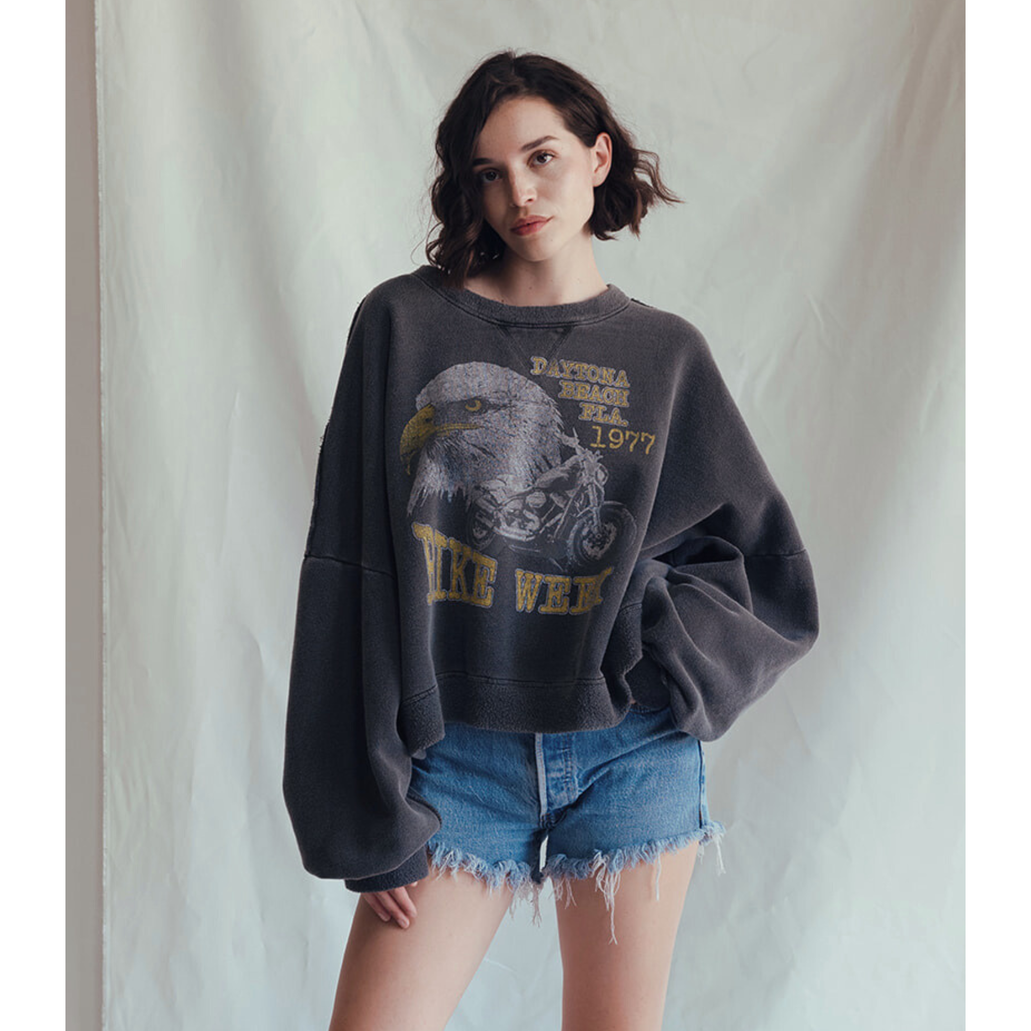A woman with wavy dark hair wears the People Of Leisure Daytona Sweatshirt, which is oversized and features a vintage eagle graphic, paired with frayed denim shorts against a softly textured light background.