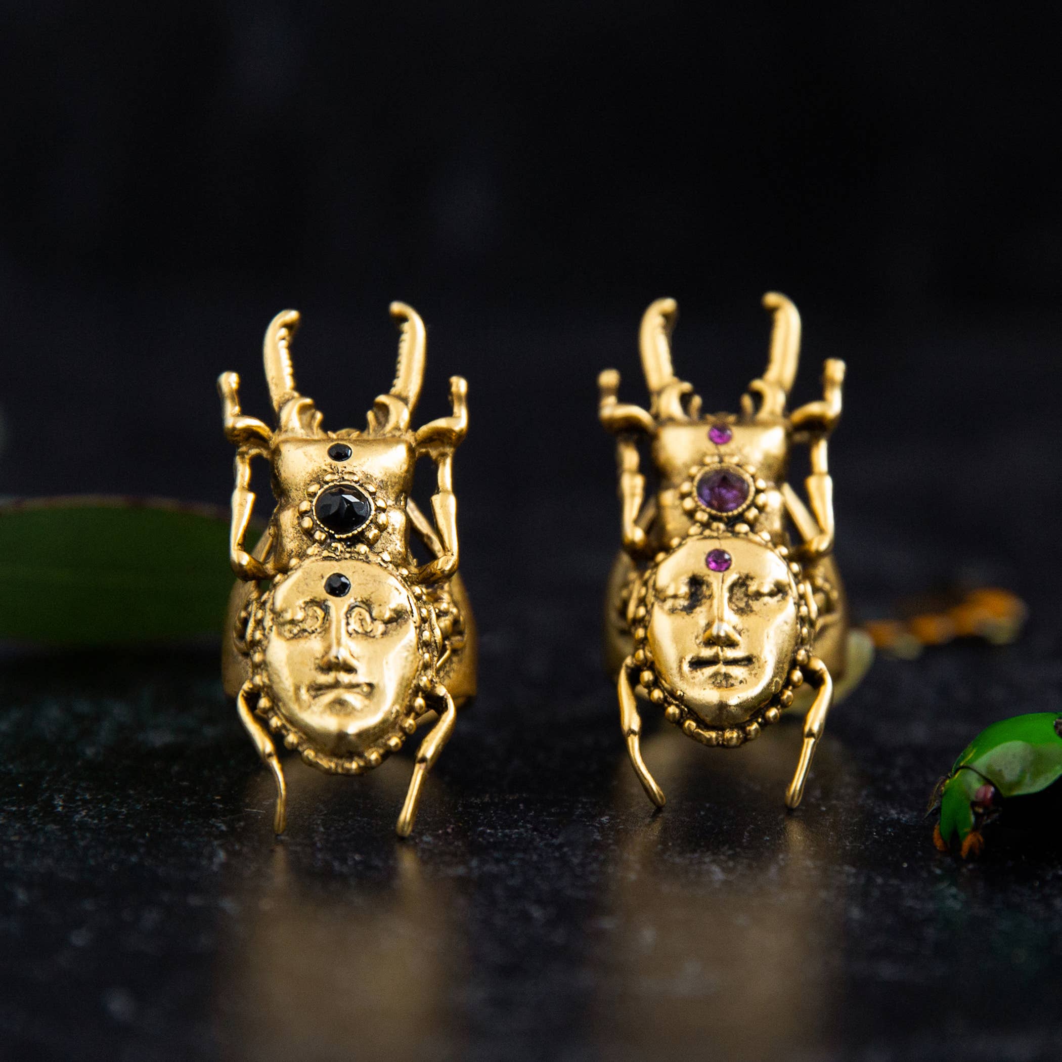 Pair of gold Hidden Magic Stag Beetle Ring pieces with detailed faces and black and purple stone accents on a dark surface