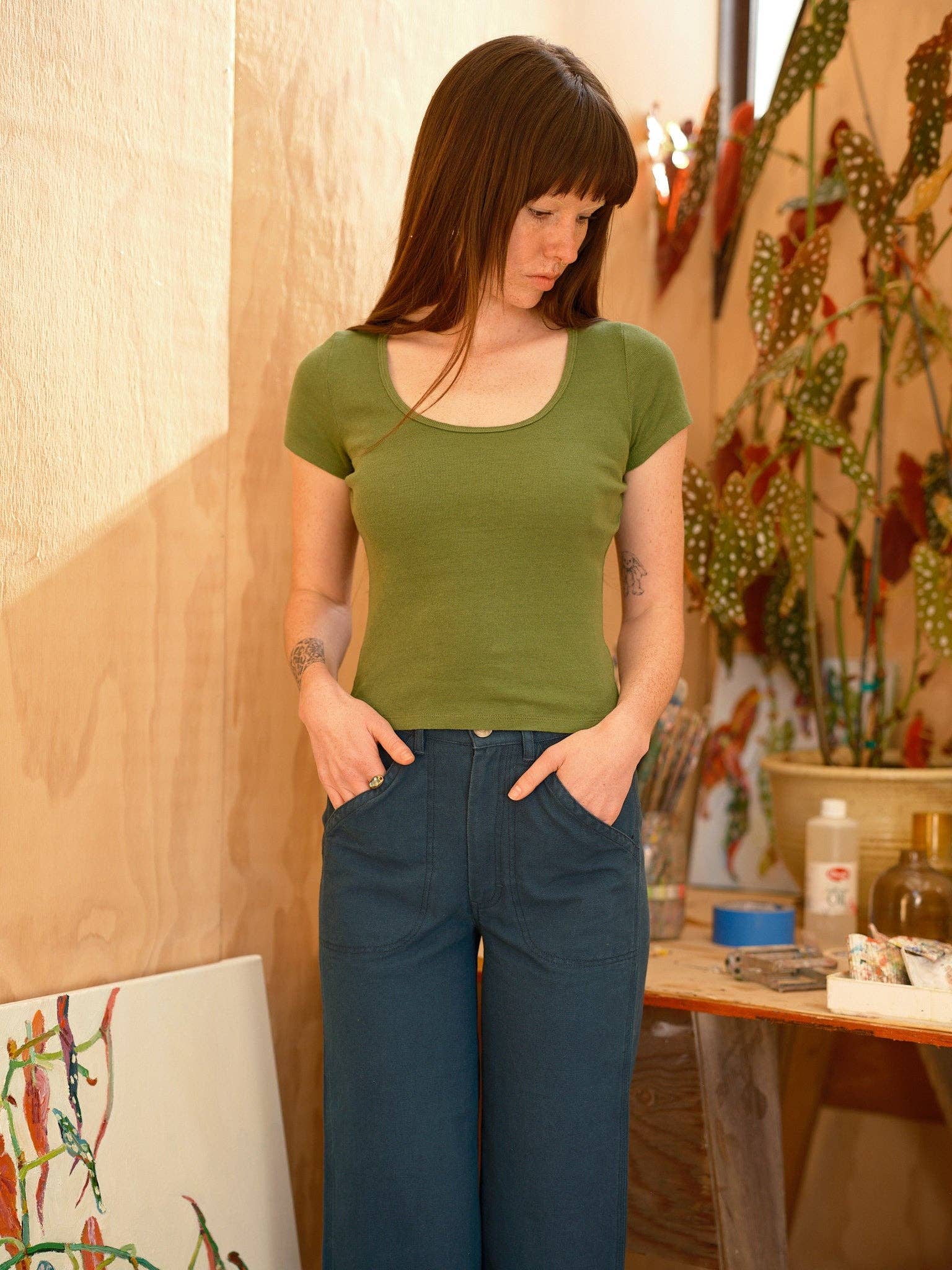 A woman with long brown hair, wearing the Mollusk Scoop Ribsy Tee in green and blue pants, stands indoors with her hands in her pockets; art supplies, plants, and a painting appear in the background.