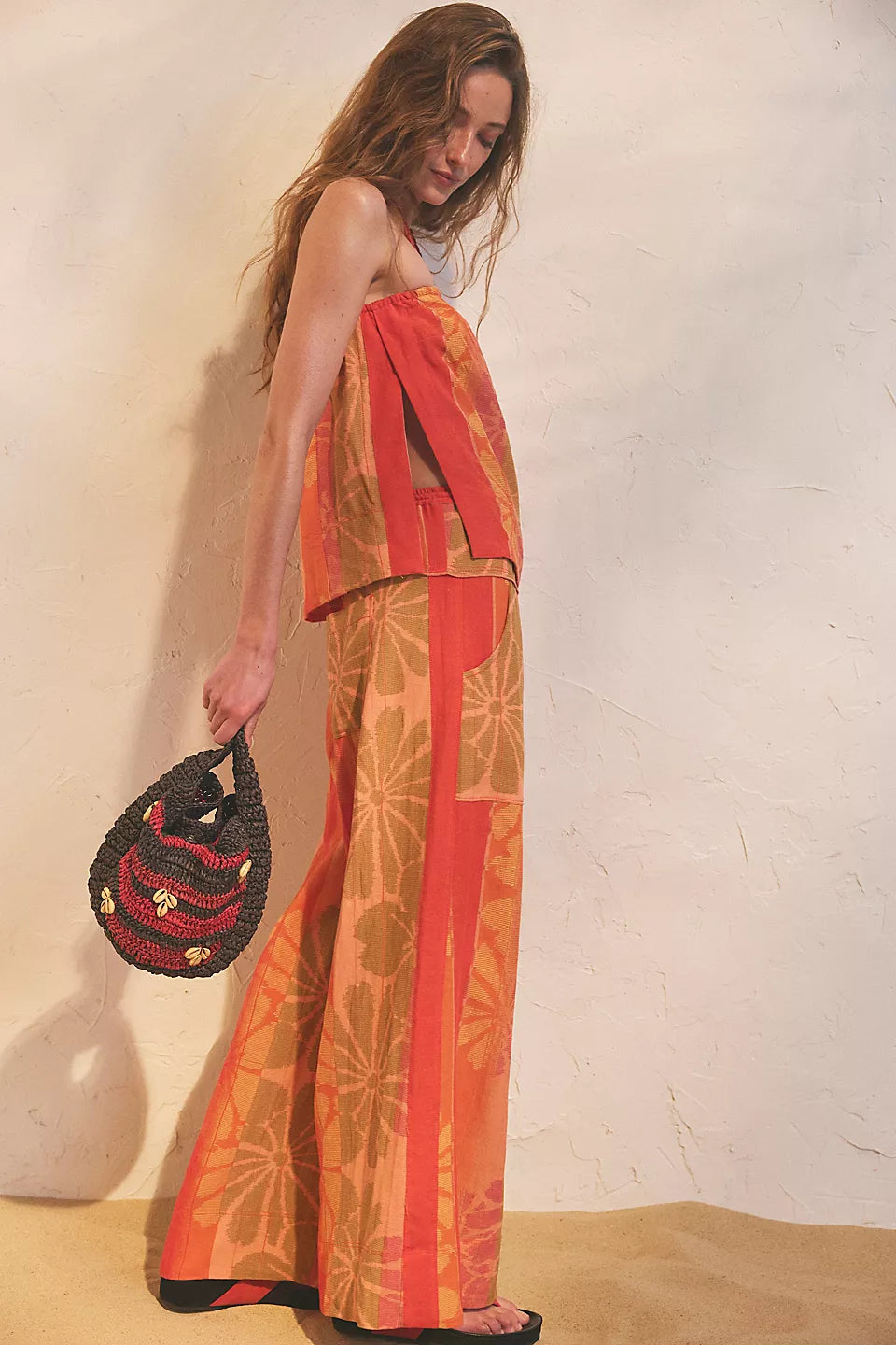 Woman wearing vibrant orange floral pants and strapless top with side slits, holding a black and red woven bag, FREE PEOPLE DEVELLA SET