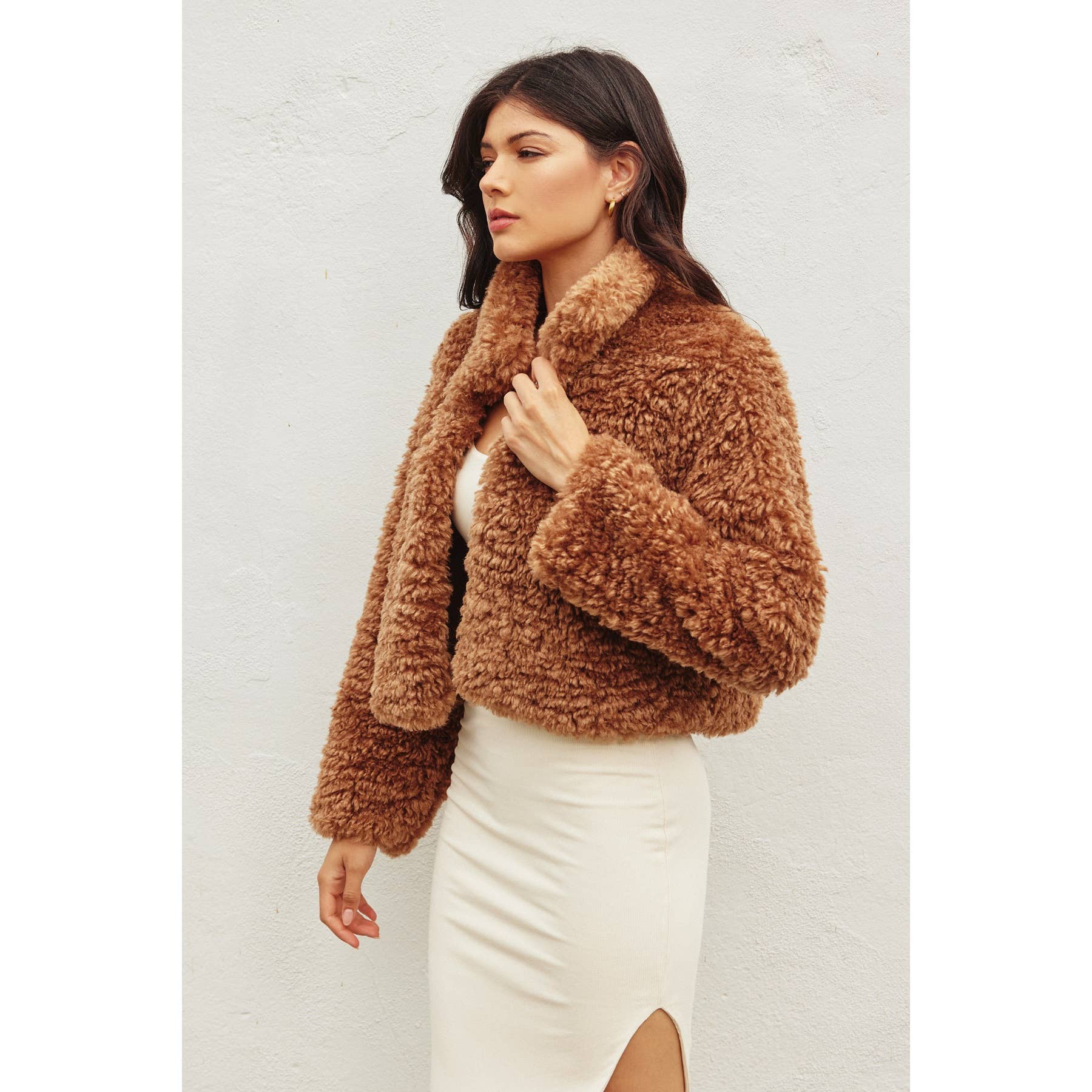 A woman with long dark hair models the Dress Forum Soft Teddy Open Front Cropped Jacket over a cream-colored dress with a thigh-high slit, posed against a plain white background.