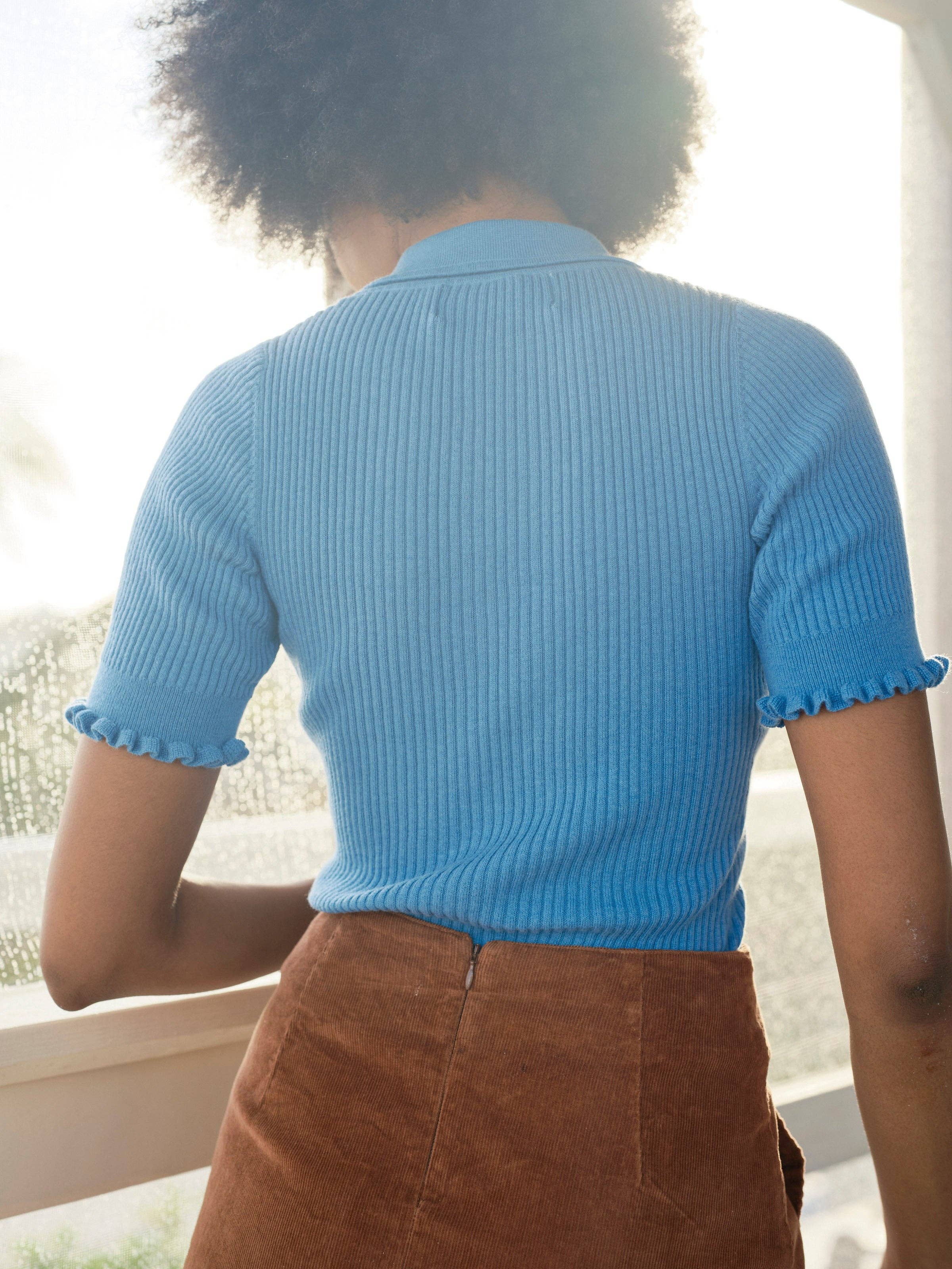 A person with curly hair stands indoors, facing away, wearing the Mollusk Belle Tee—a vintage-inspired ribbed blue short-sleeve top—paired with a high-waisted brown skirt as sunlight streams in through a window.