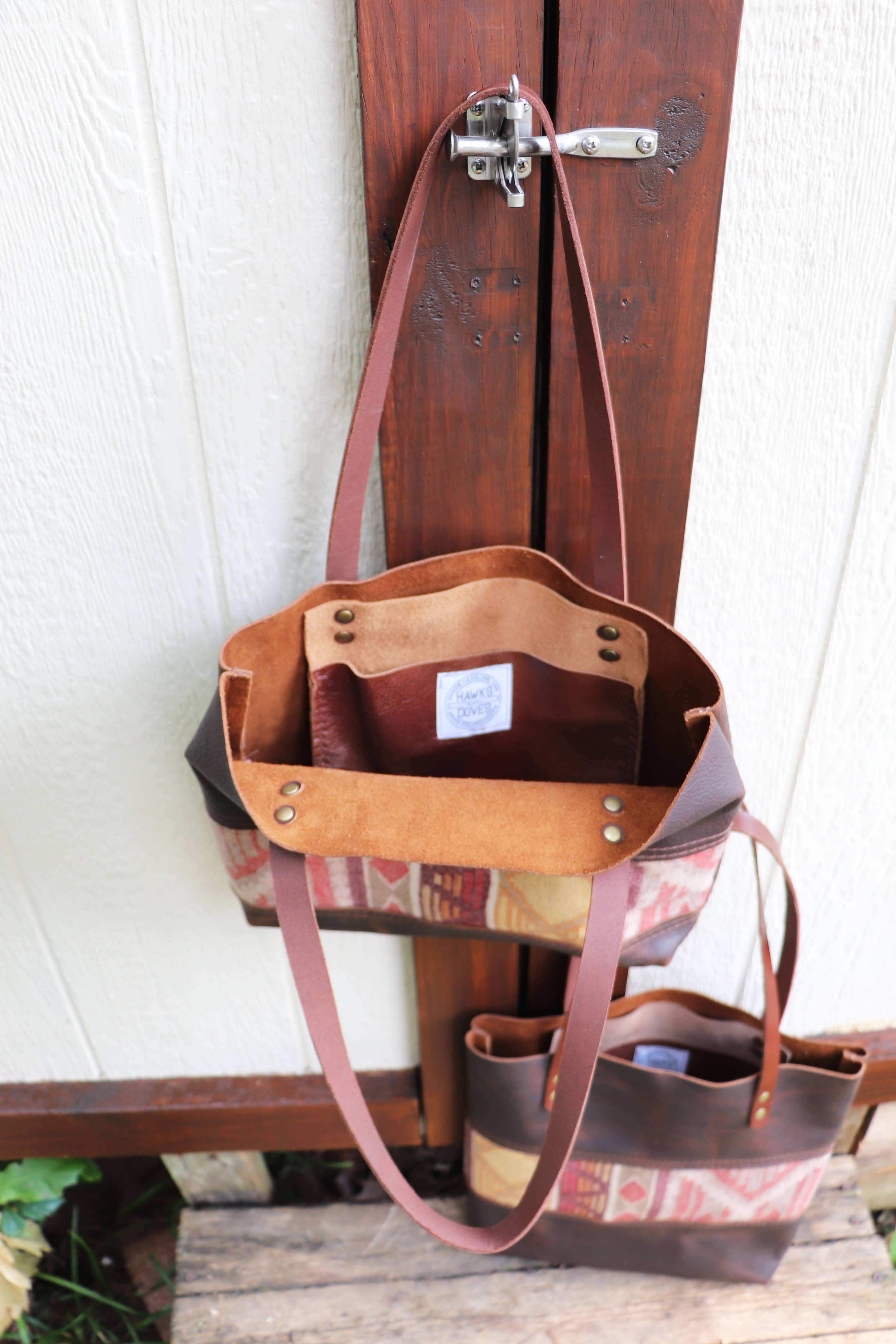 A Hawks & Doves Wool Tote Short, crafted from full grain leather and patterned wool with brown straps, hangs open by a metal latch on a wooden door. A similar bag rests on a wooden surface below.