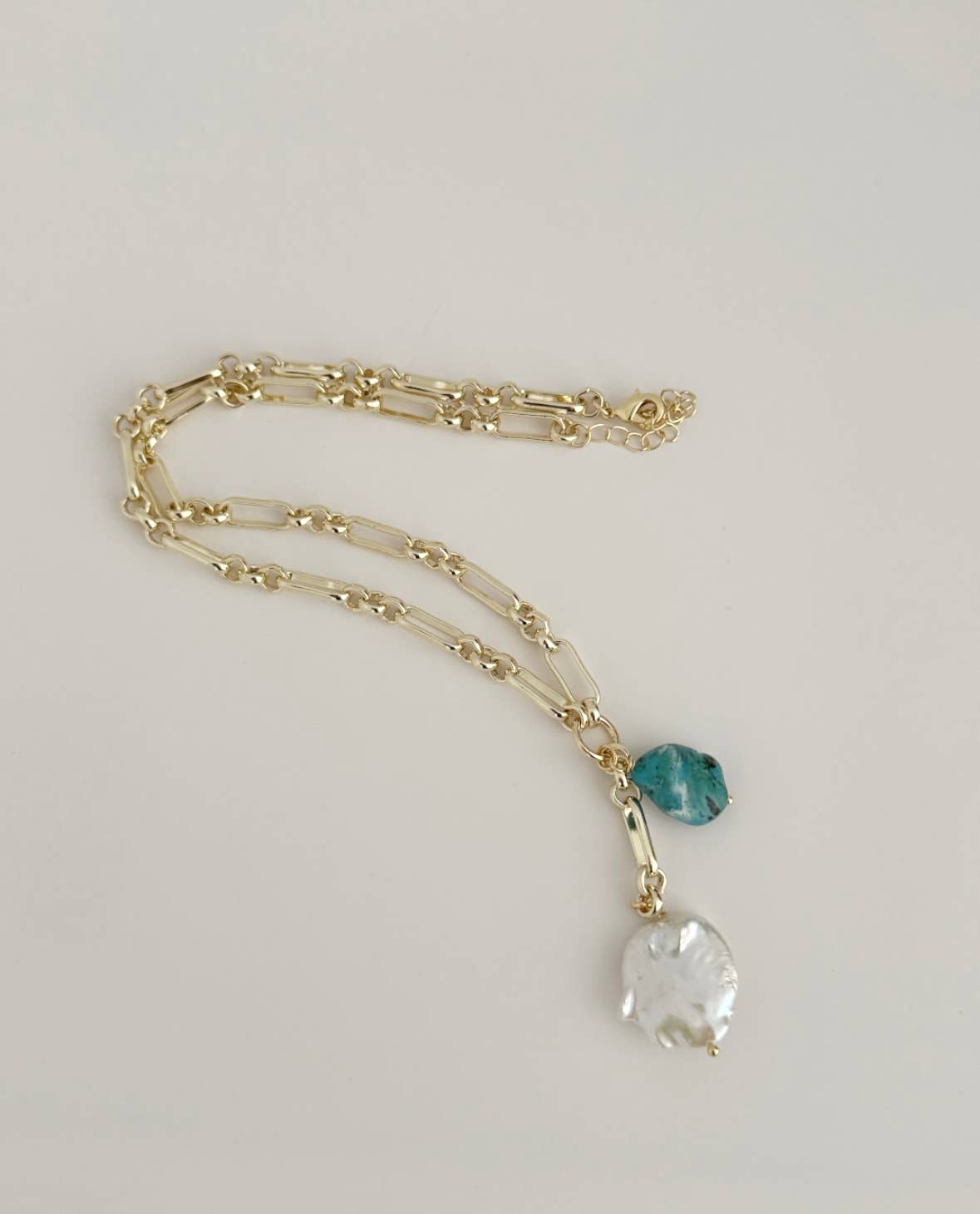 The Azure Tide Necklace by Tramps+Thieves features a gold chain with two irregular pendants—a baroque pearl and a teal translucent blue stone—beautifully displayed against a plain light background.