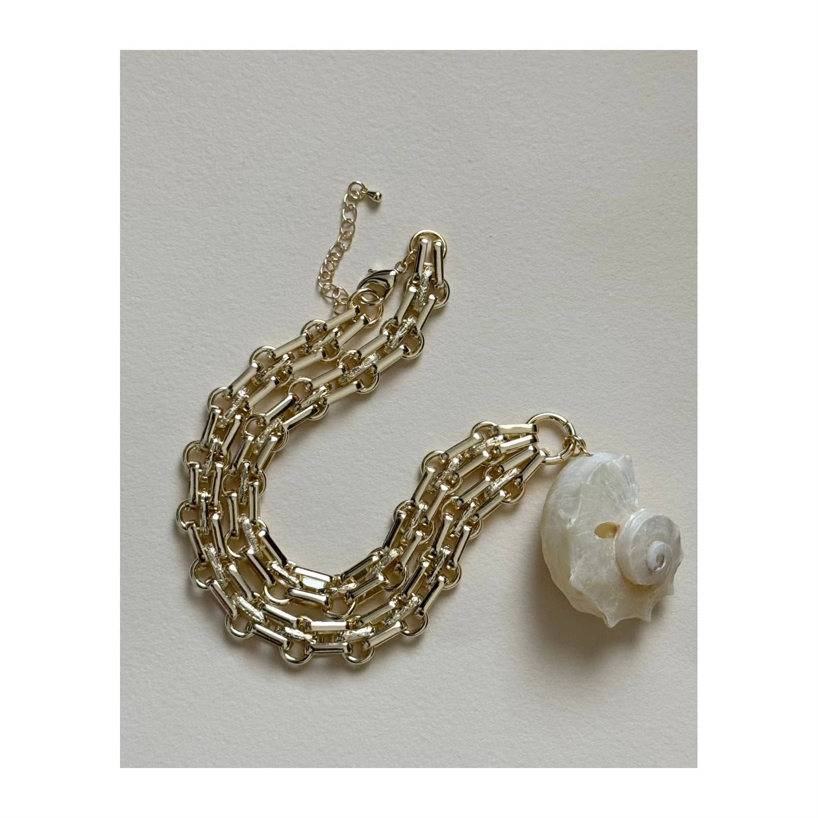 The Golden Nautilus Necklace by Tramps+Thieves features three rows of gold-filled chains and a striking white seashell charm, perfect for layering with your favorite coastal jewelry and displayed on a light beige surface.
