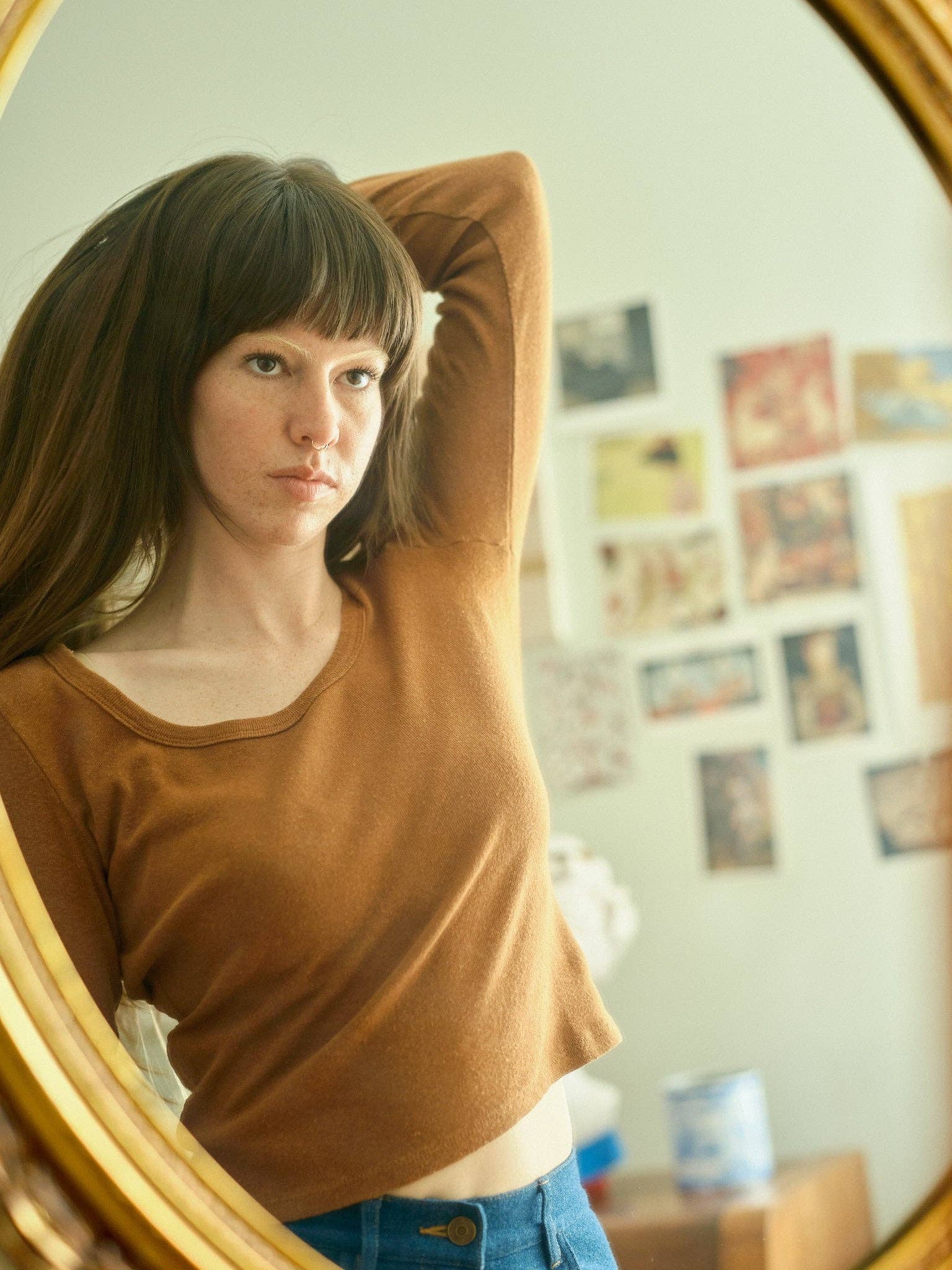 A woman with long brown hair and bangs gazes at her reflection in a round mirror, wearing the Mollusk Long Sleeve Rib Tee and blue pants. Art and photos decorate the wall behind her.