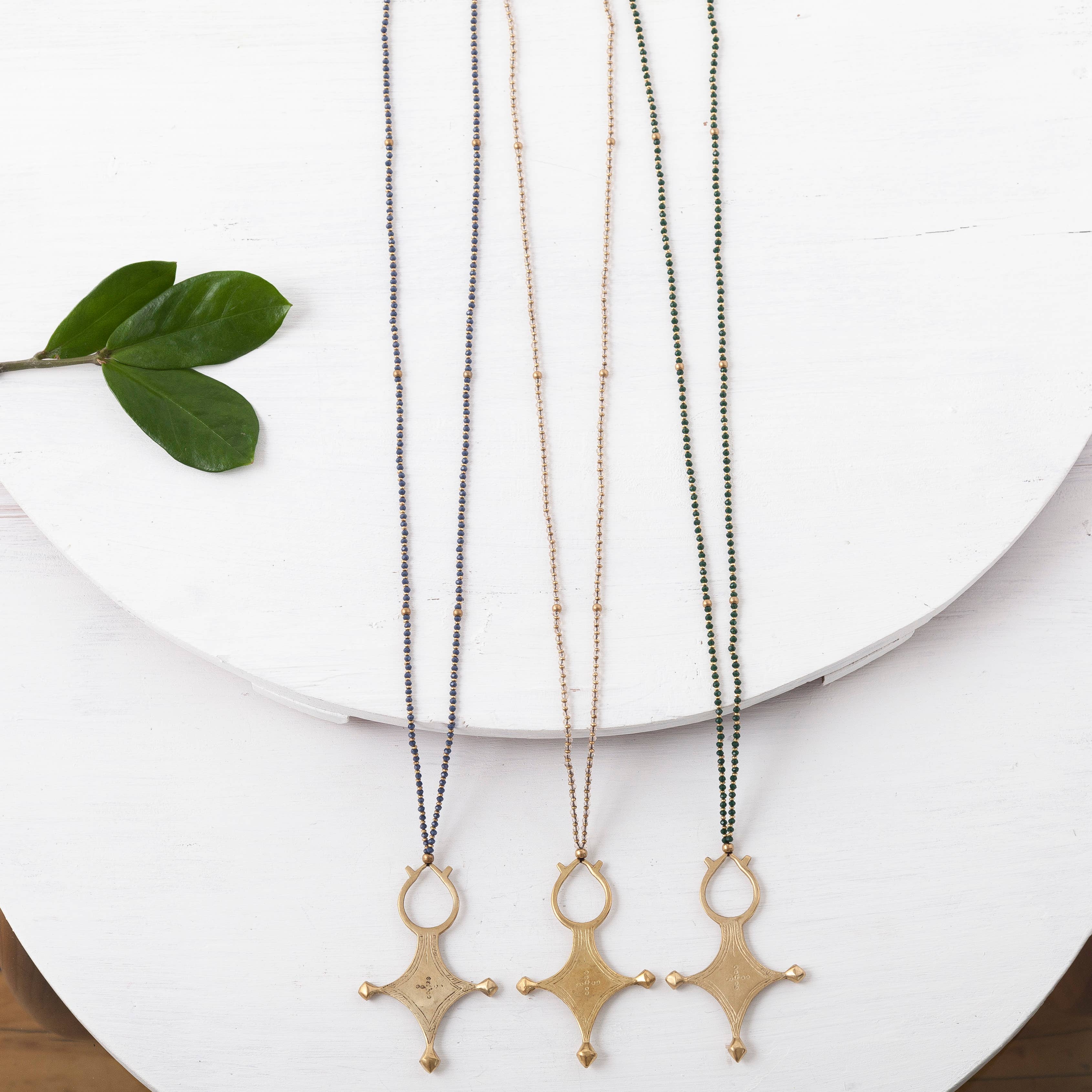 Three long necklaces, including Baizaar's Color Pop Beaded Tribal Cross Necklace, are shown on a round white surface with green leaves. Each gold-toned pendant hangs from a distinct beaded chain.