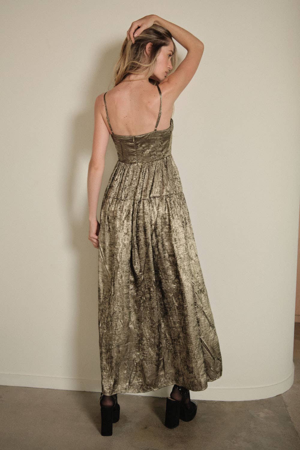 A woman faces a wall with her back to the camera, wearing the Promesa USA Textured Metallic Fit-and-Flare Corset Maxi Dress with thin straps and black platform shoes. The dress has a fit-and-flare shape, and she raises one hand to touch her head.