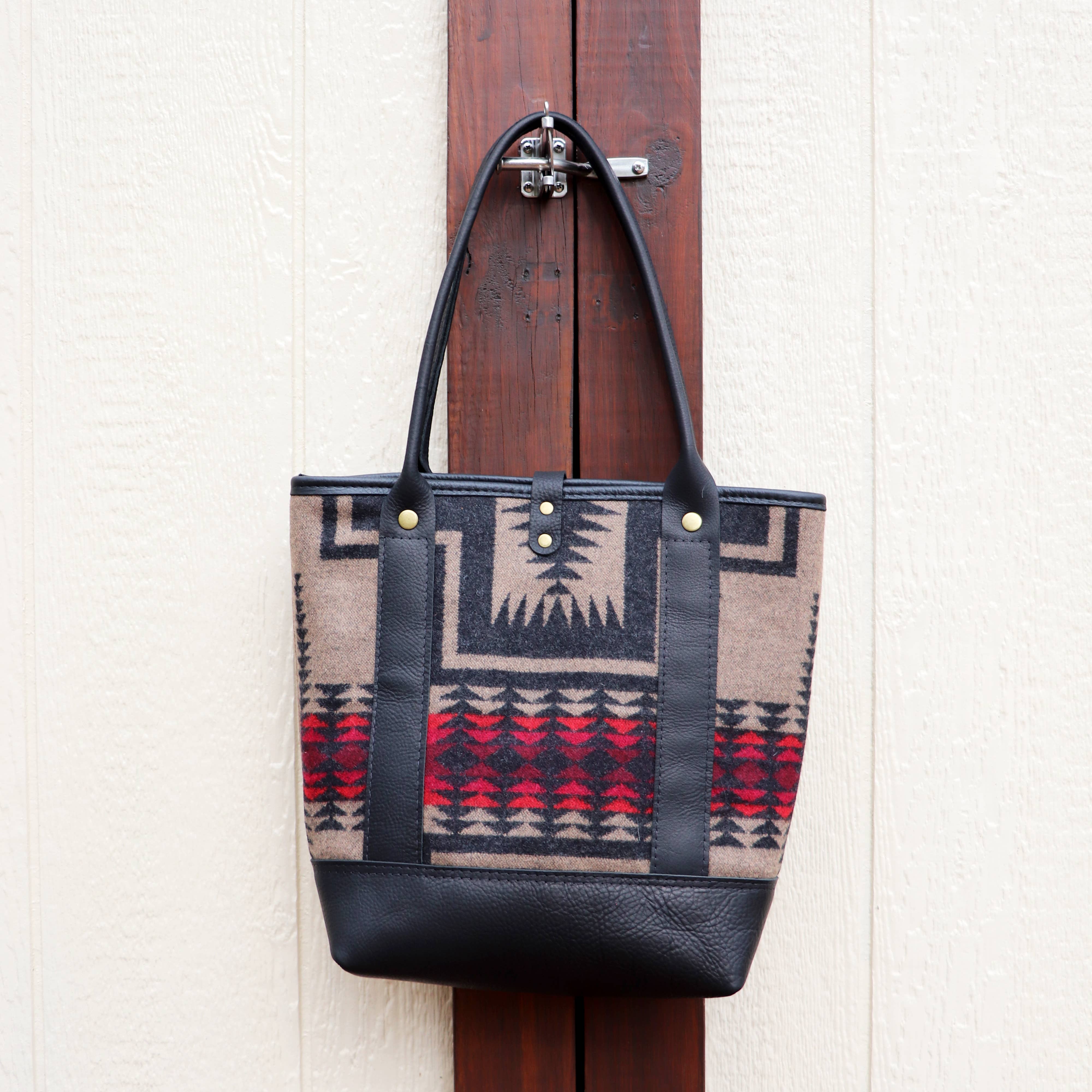 The Pearl Wool and Leather Tote by Hawks & Doves, handmade in Raleigh, features a beige, gray, black, and red geometric Pendleton wool body with black full grain leather handles and base. Displayed on a wooden latch against a textured wall.