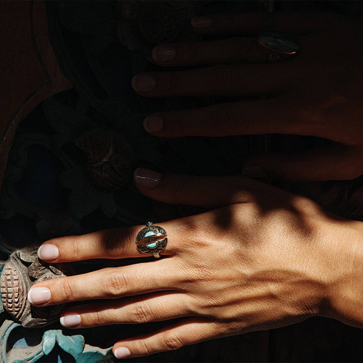 Two hands with manicured nails rest on carved wood, adorned with the bahgsu jewels Natural Mystic Ring - Turquoise. Sunlight plays across the ring, accentuating its vibrant hue and the natural patterns of the surface below.
