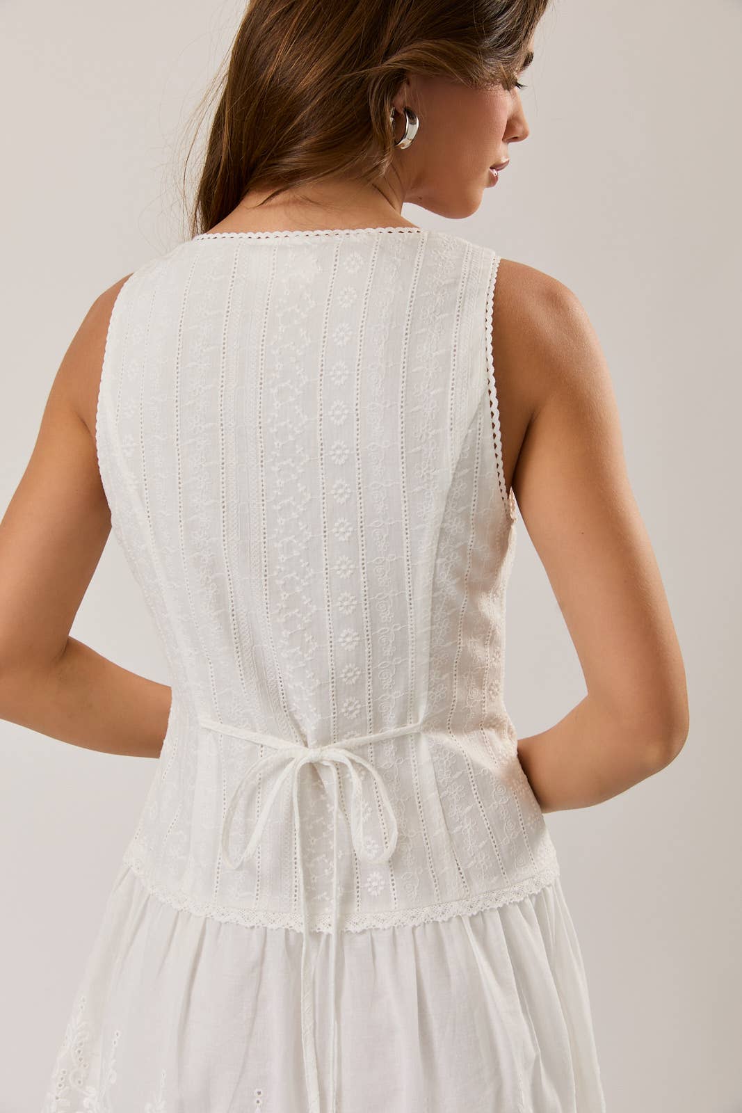 Woman wearing a white Sleeveless Eyelet Mini Dress featuring delicate embroidery and a tie-back waist detail.