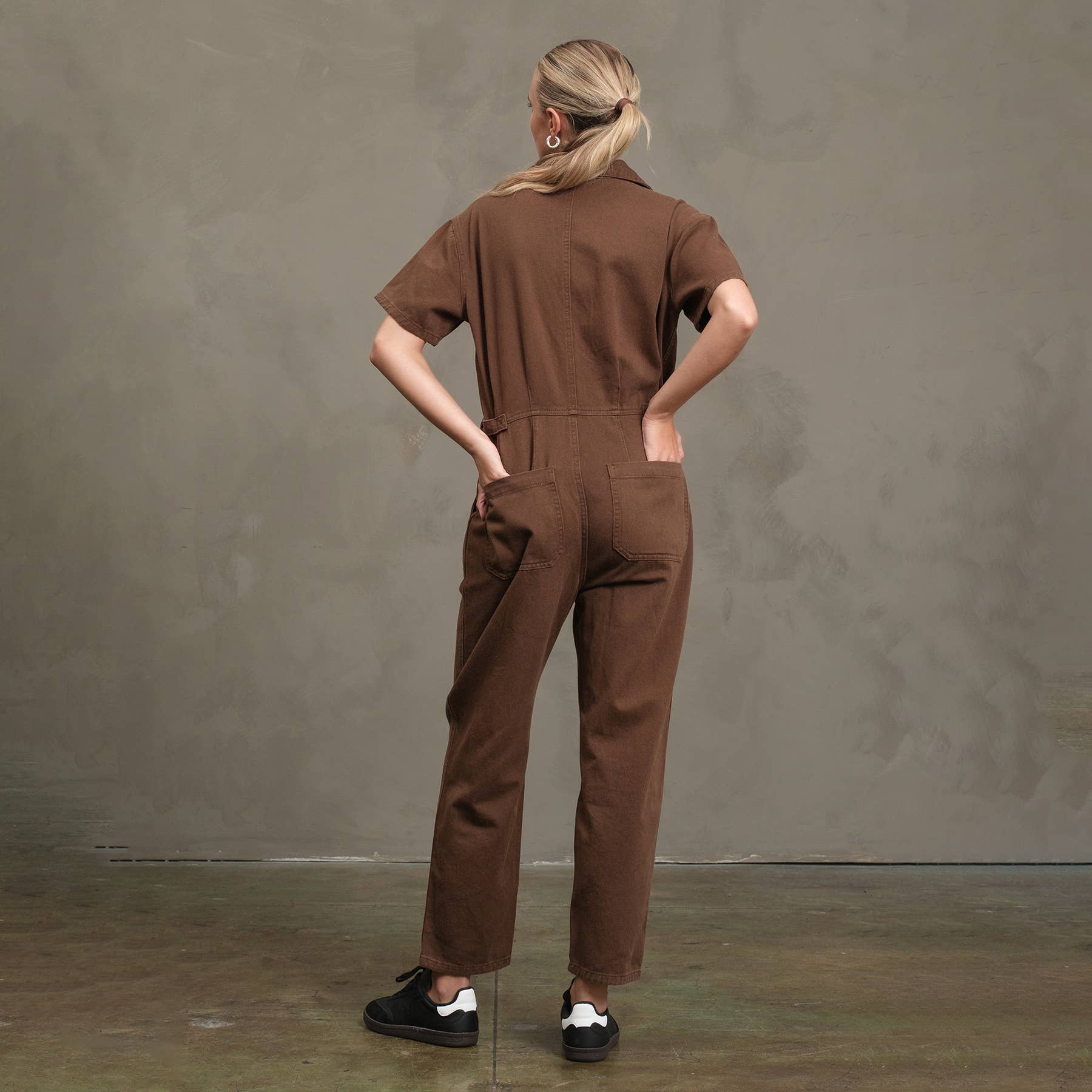 A person with blond hair in a ponytail stands facing away against a gray backdrop, wearing the RAISONNEL Field Jumpsuit - RJP1001, made from breathable cotton, paired with black sneakers and one hand in a back pocket.