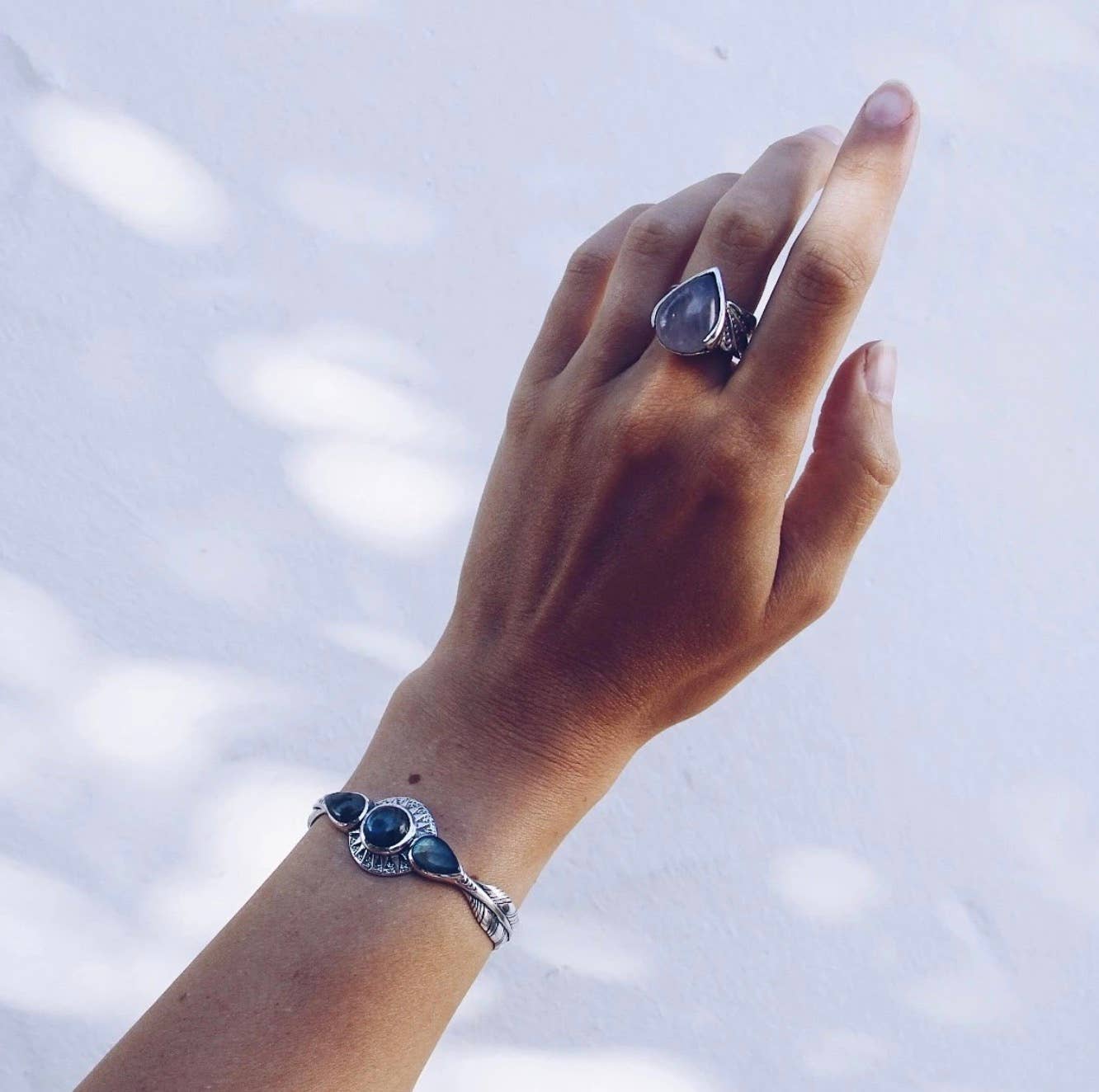 A hand wearing the bahgsu jewels Winged Ring, a silver ring with a large blue stone, paired with a matching silver bracelet adorned with blue stones, shown against a softly lit white background.