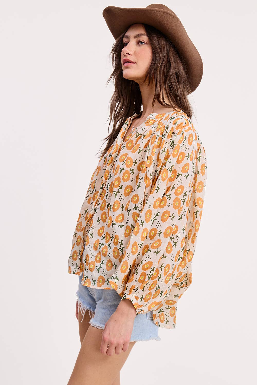 A woman in an Ultramarine Meadow Breeze Linen Blouse, featuring yellow and orange floral prints, wears a wide-brim brown hat and light blue denim shorts while standing sideways against a plain white background.