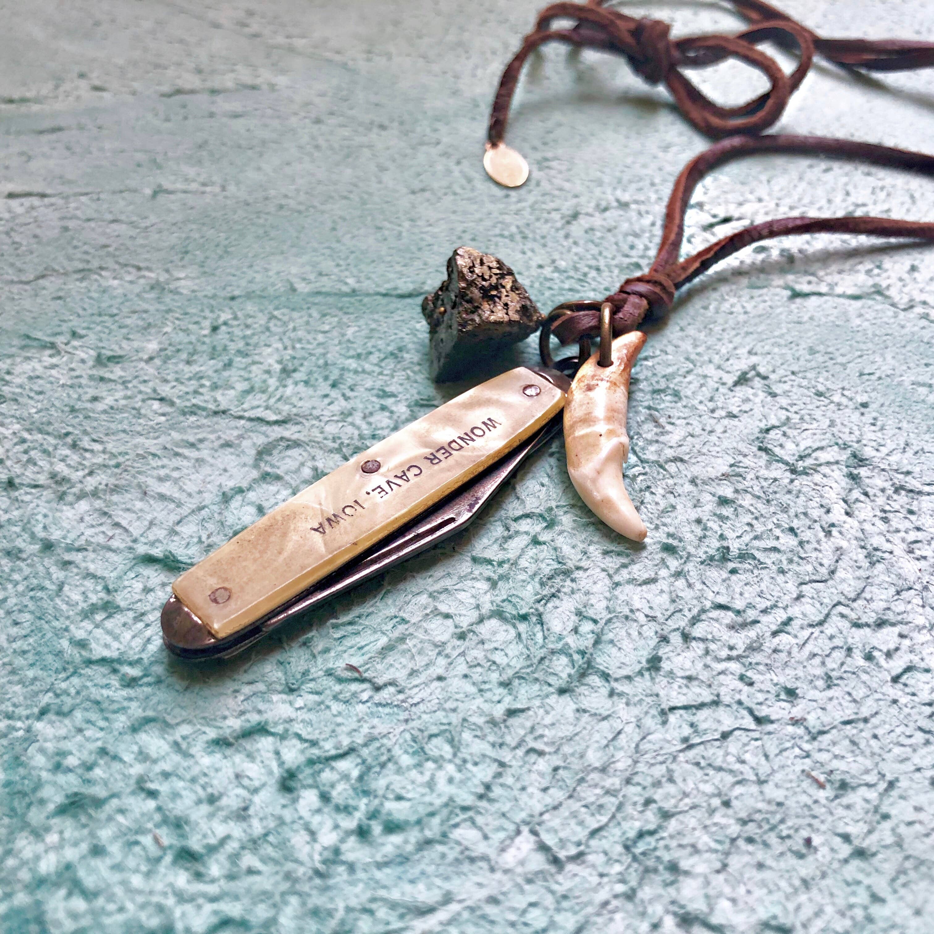 A close-up of the ASTALI Vintage Pocket Knife & Coyote Tooth Necklace, featuring a pyrite pendant and deerskin leather cord, displayed on a textured turquoise surface.
