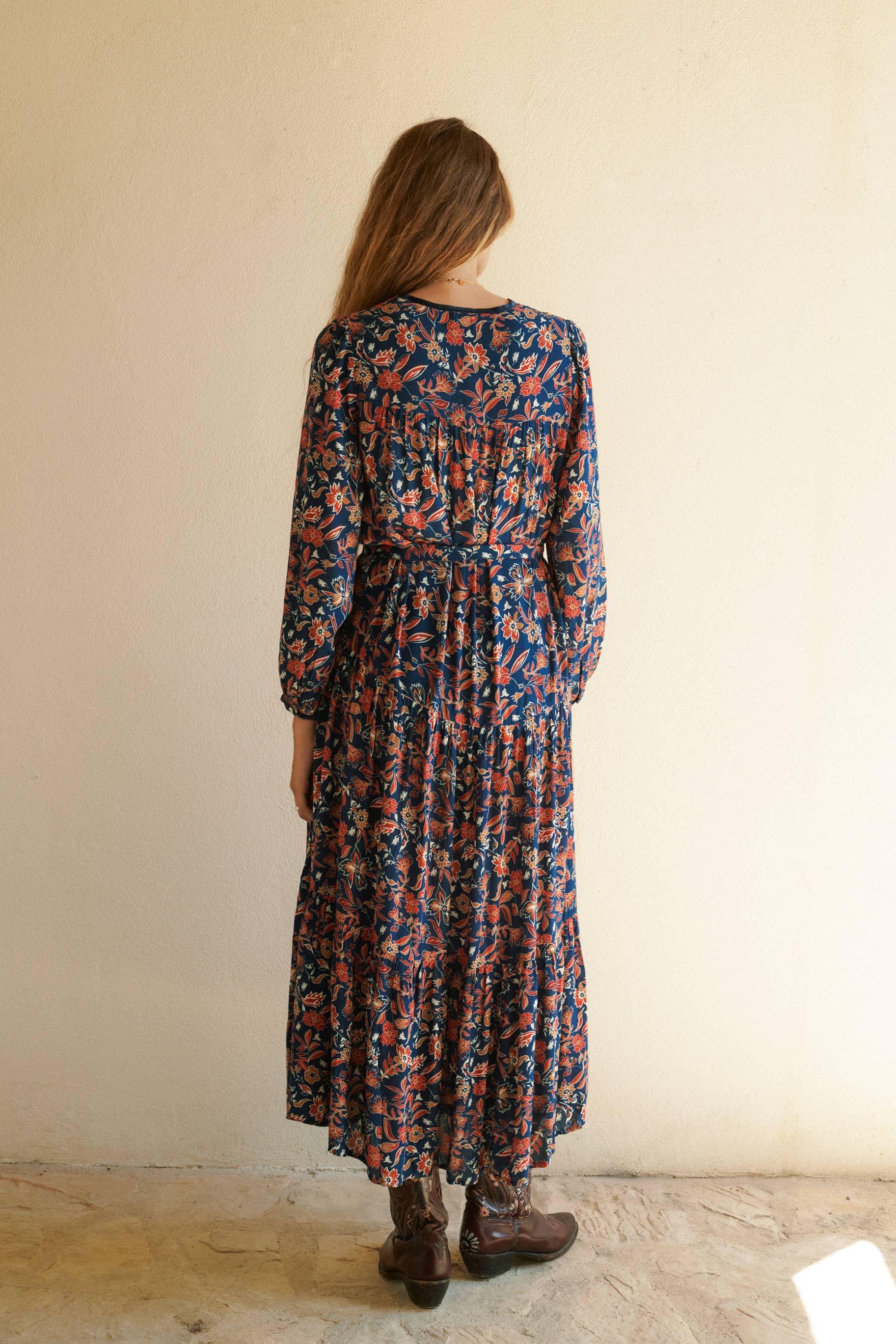 A woman with long brown hair stands facing a cream-colored wall, wearing the Aura Dress Navy by Sonmer Shop USA. Her back is to the camera, arms at her sides, highlighting the bohemian maxi dress style.