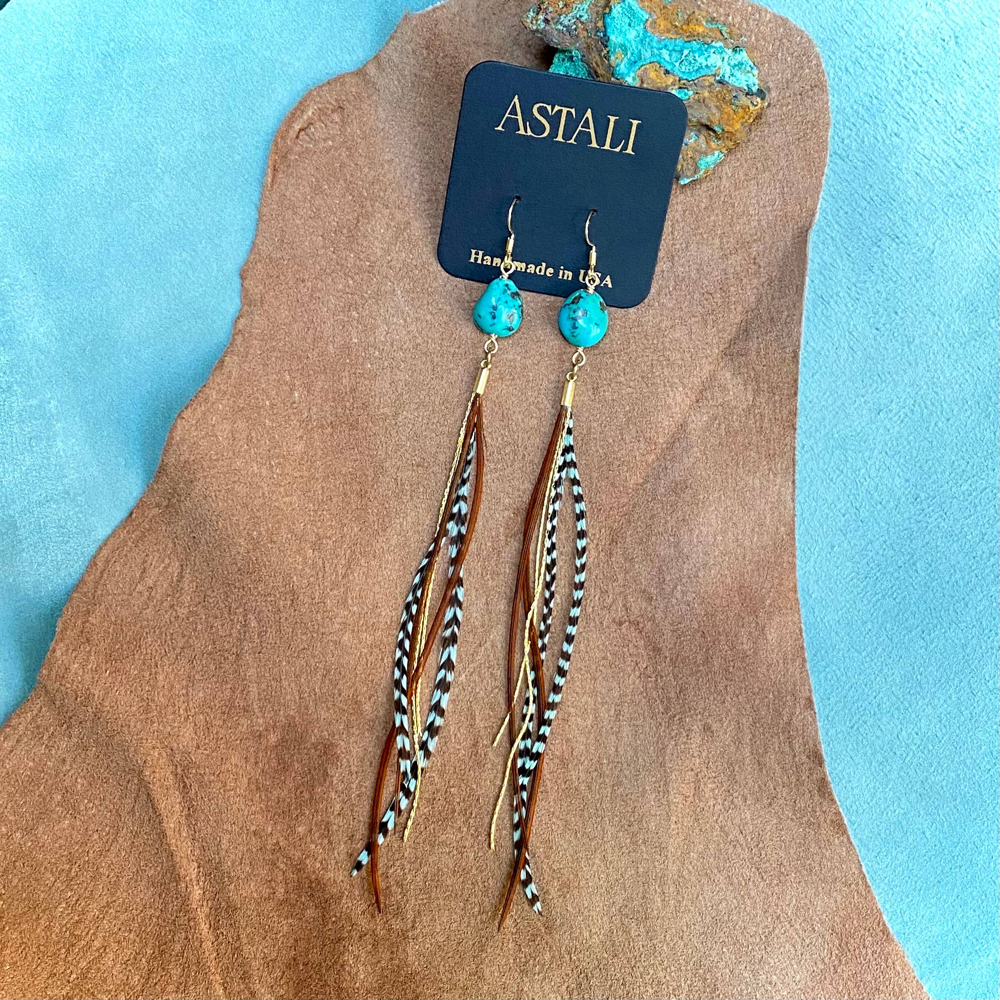 The ASTALI Turquoise & Mini Feather Earrings feature turquoise stone tops, gold hooks, and teal grizzly and fawn feathers, displayed on a branded card against a tan and blue background.