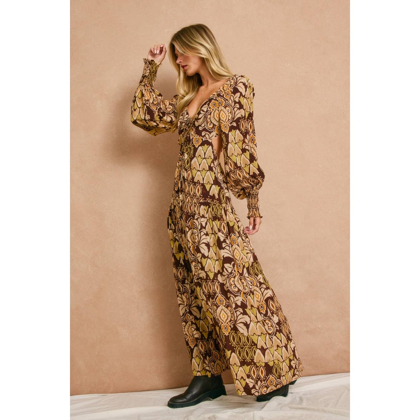 A woman wears the Dress Forum Stone Ember Keyhole Long Sleeve Maxi Dress and black ankle boots, standing sideways against a beige wall with one arm raised to her head.