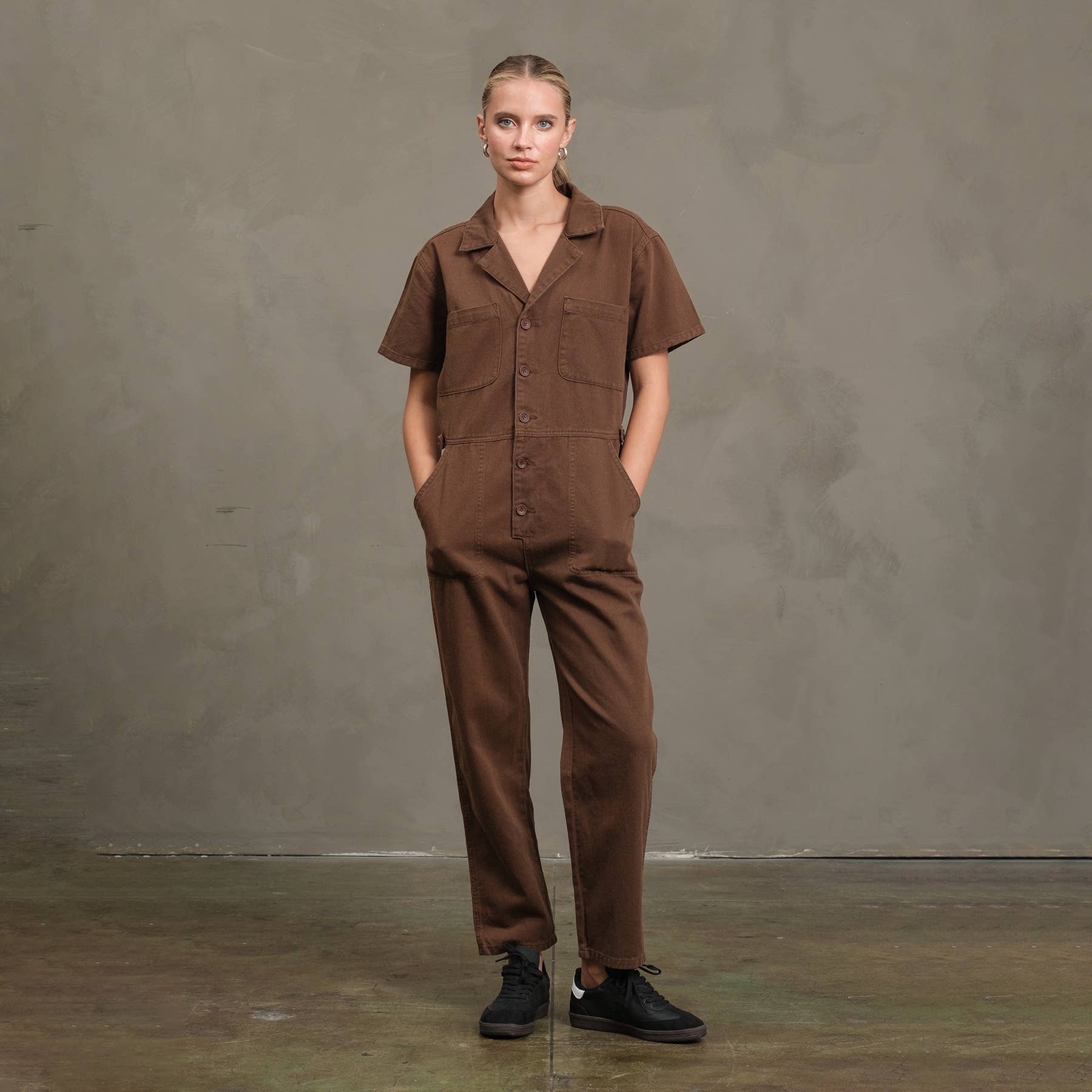 A person stands against a plain gray wall, wearing the RAISONNEL Field Jumpsuit - RJP1001 in brown cotton and black sneakers, with hands in pockets, looking directly at the camera.