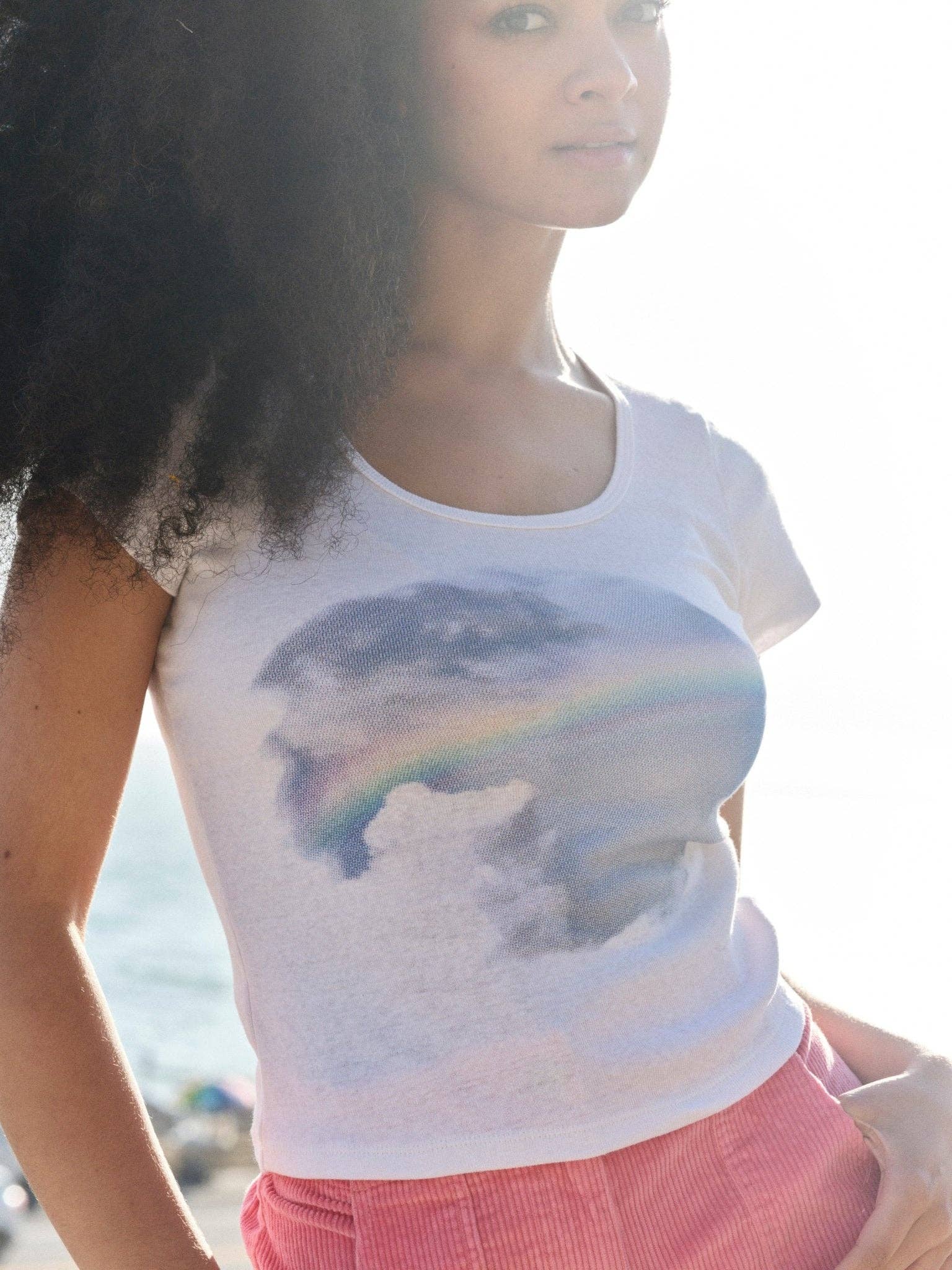 A young woman with voluminous curly hair wears the Mollusk CloudBreak Tee, made from sustainable hemp cotton. She stands in bright sunlight, with the California surf culture coastline softly blurred behind her.