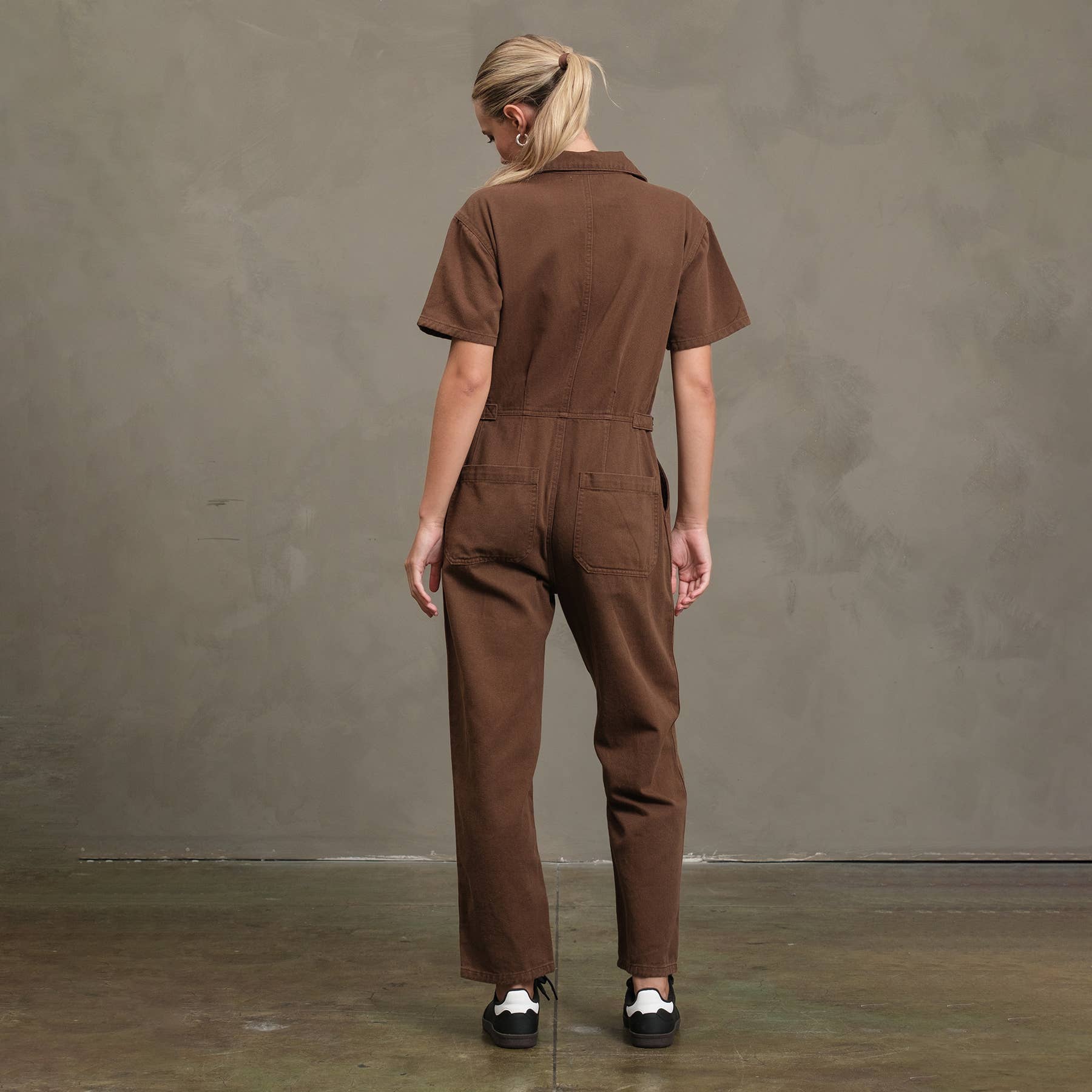 A person with blonde hair in a ponytail faces away, wearing the RAISONNEL Field Jumpsuit (RJP1001)—a short-sleeved, breathable brown cotton utility jumpsuit—and black-and-white sneakers, set against a textured gray wall and concrete floor.